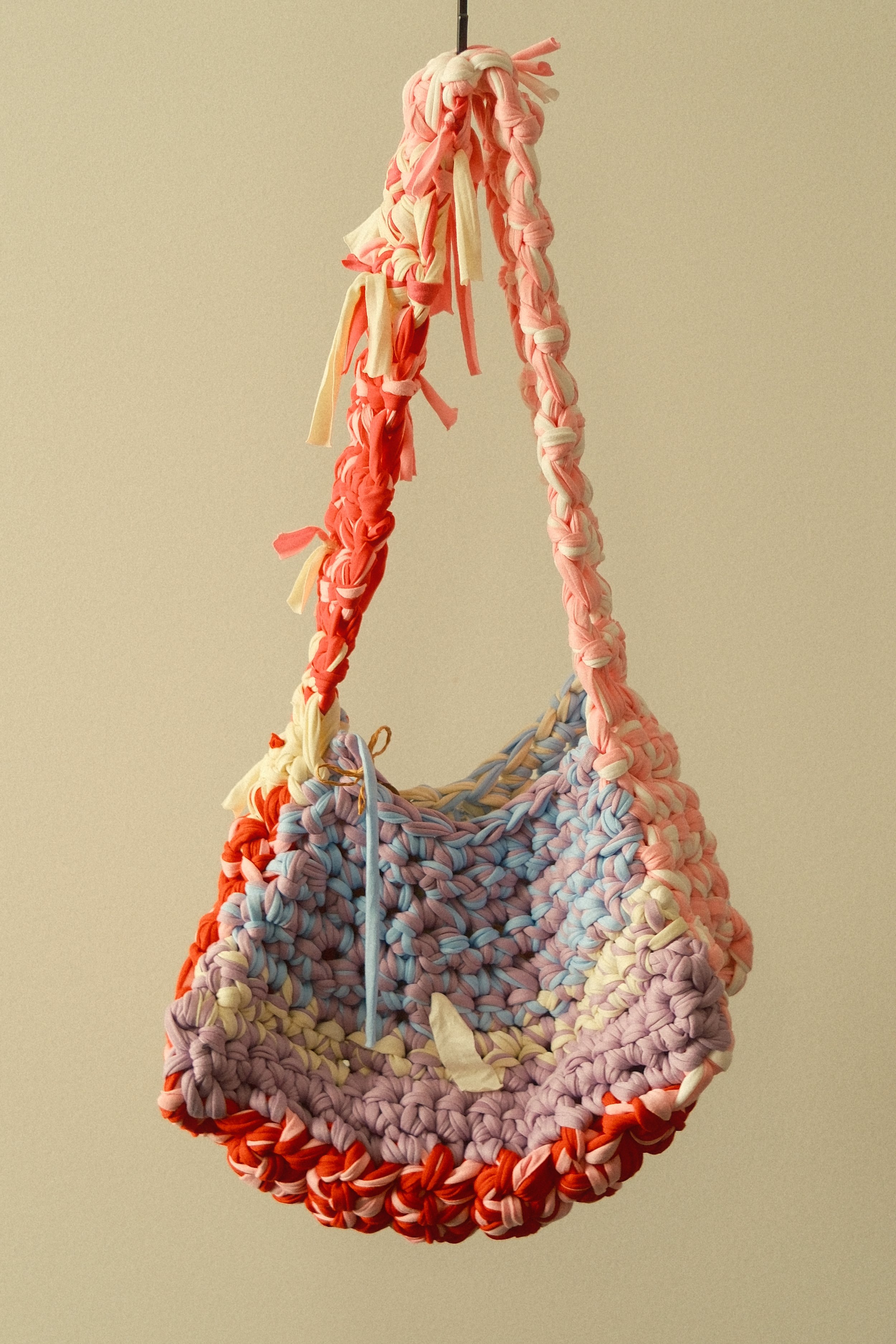 A circular purse made from multicolored interwoven fabric strips, featuring shades of pink, lavender, cream, and red, hanging against a plain beige background.