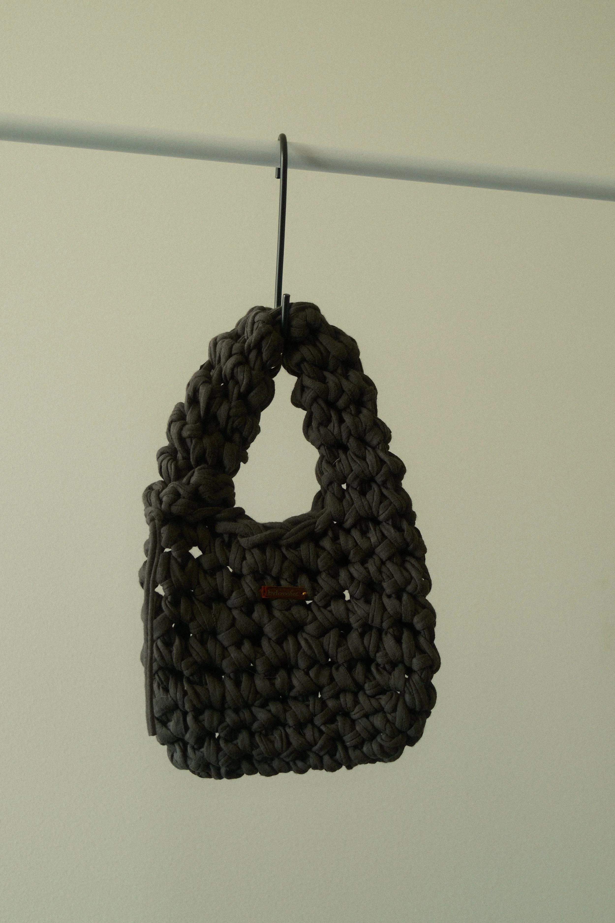 A dark gray, chunky, knit bag with a small orange tag, hanging from a black hook on a white wall.