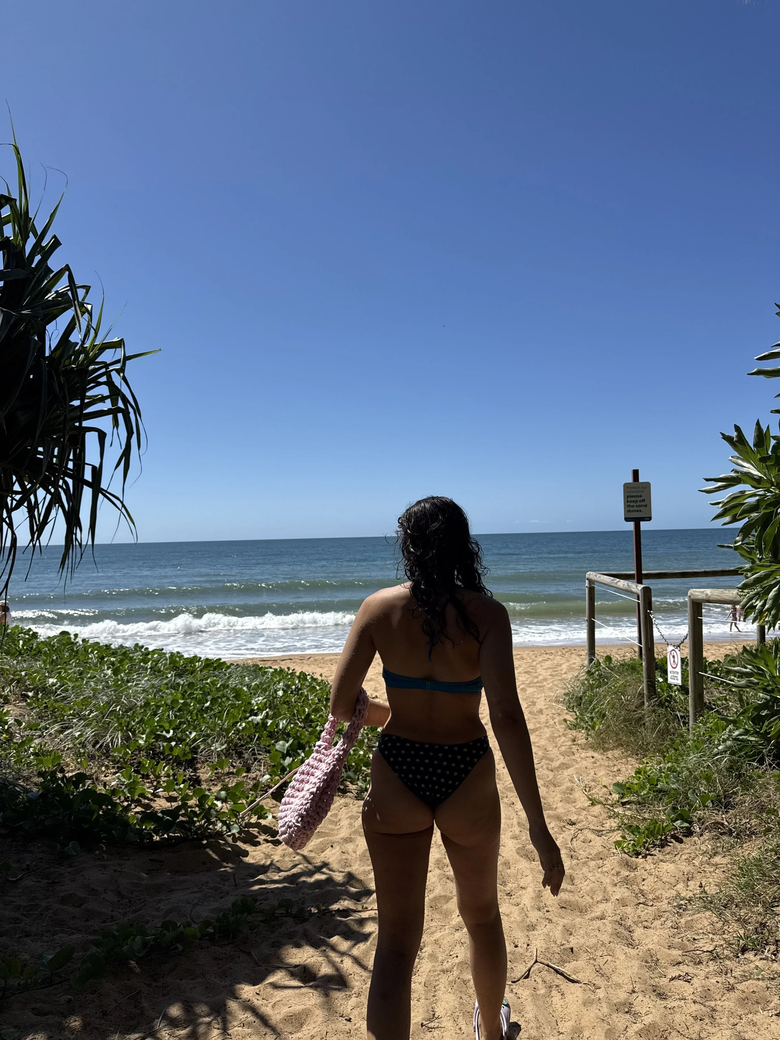 Woman in a swimsuit carrying a pink bag walking towards the beach under a blue sky, with lush green plants on either side.