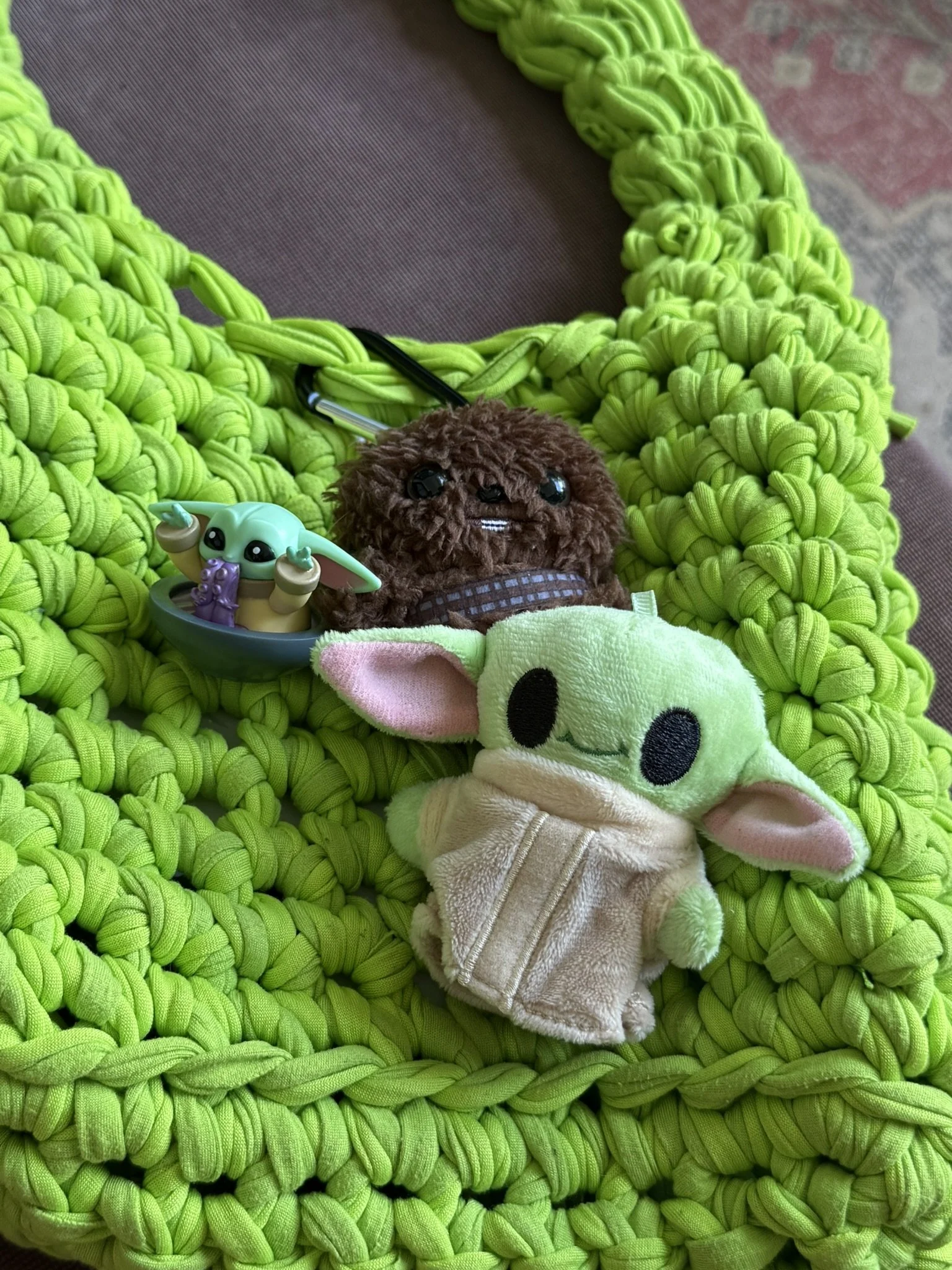 Close-up of a lime green crocheted necklace, two plush toys, one resembling Baby Yoda in a bowl and another resembling a cute koala, on a textured purple surface.