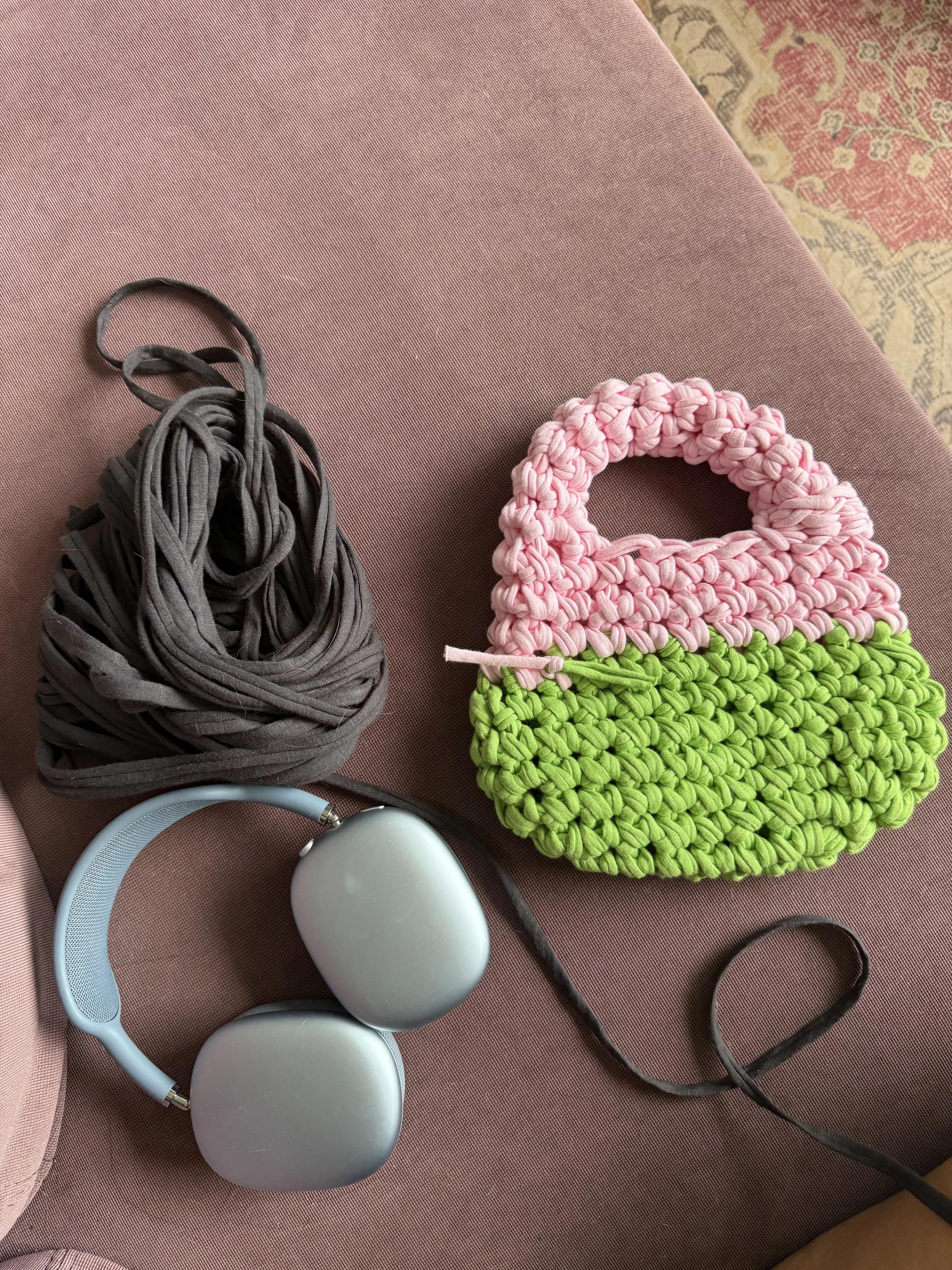 A pink textured surface with a bundle of gray fabric strips, a white headband, a white oval-shaped object, a pink and green woven bag, a white speaker, and a black hair elastic.