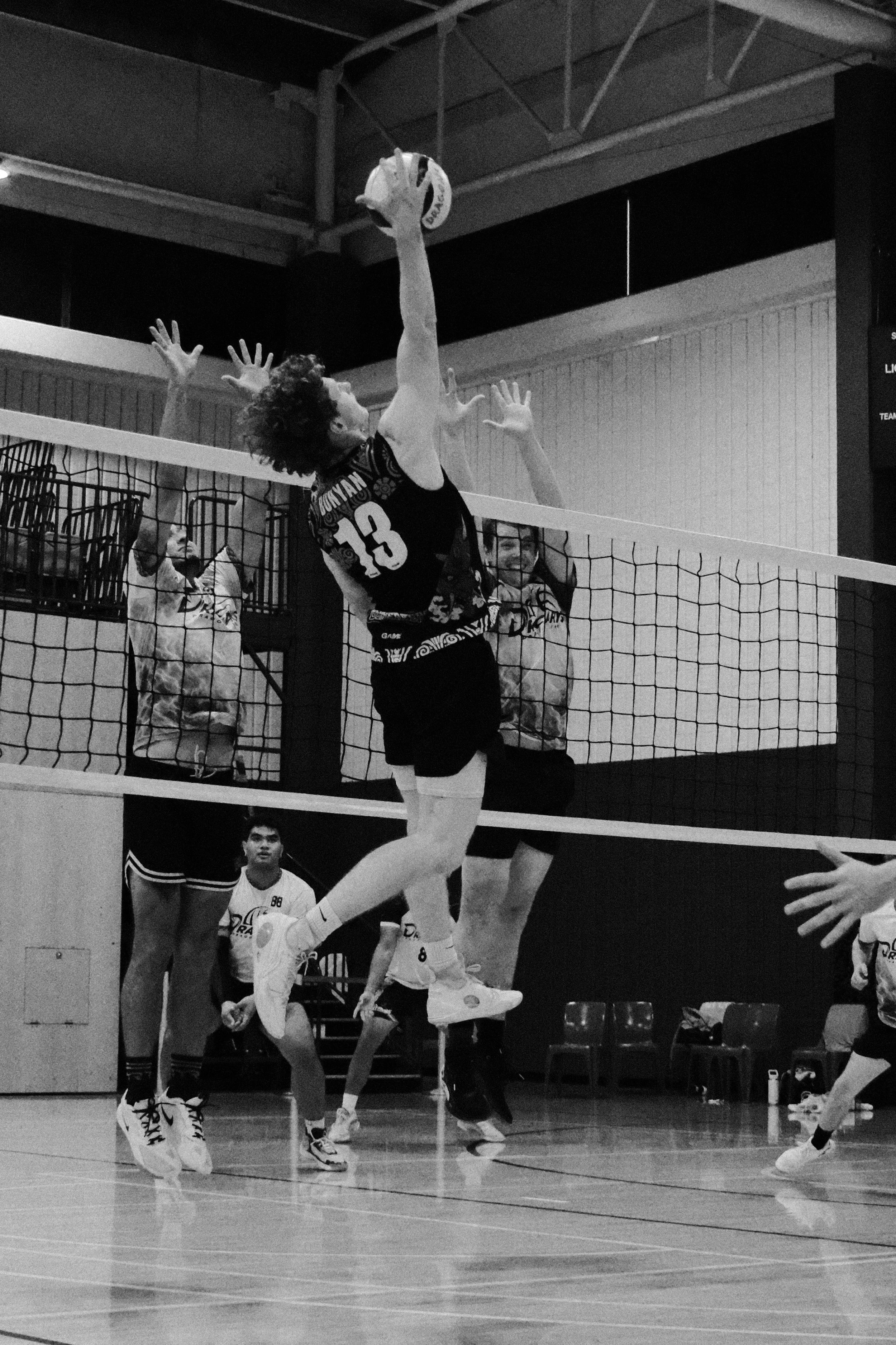 A black and white photo of a volleyball game. A player wearing a jersey with the number 13 is jumping to hit the ball over the net. Two other players and a coach or referee are visible in the background, watching the game. The scene appears to be in an indoor gymnasium.