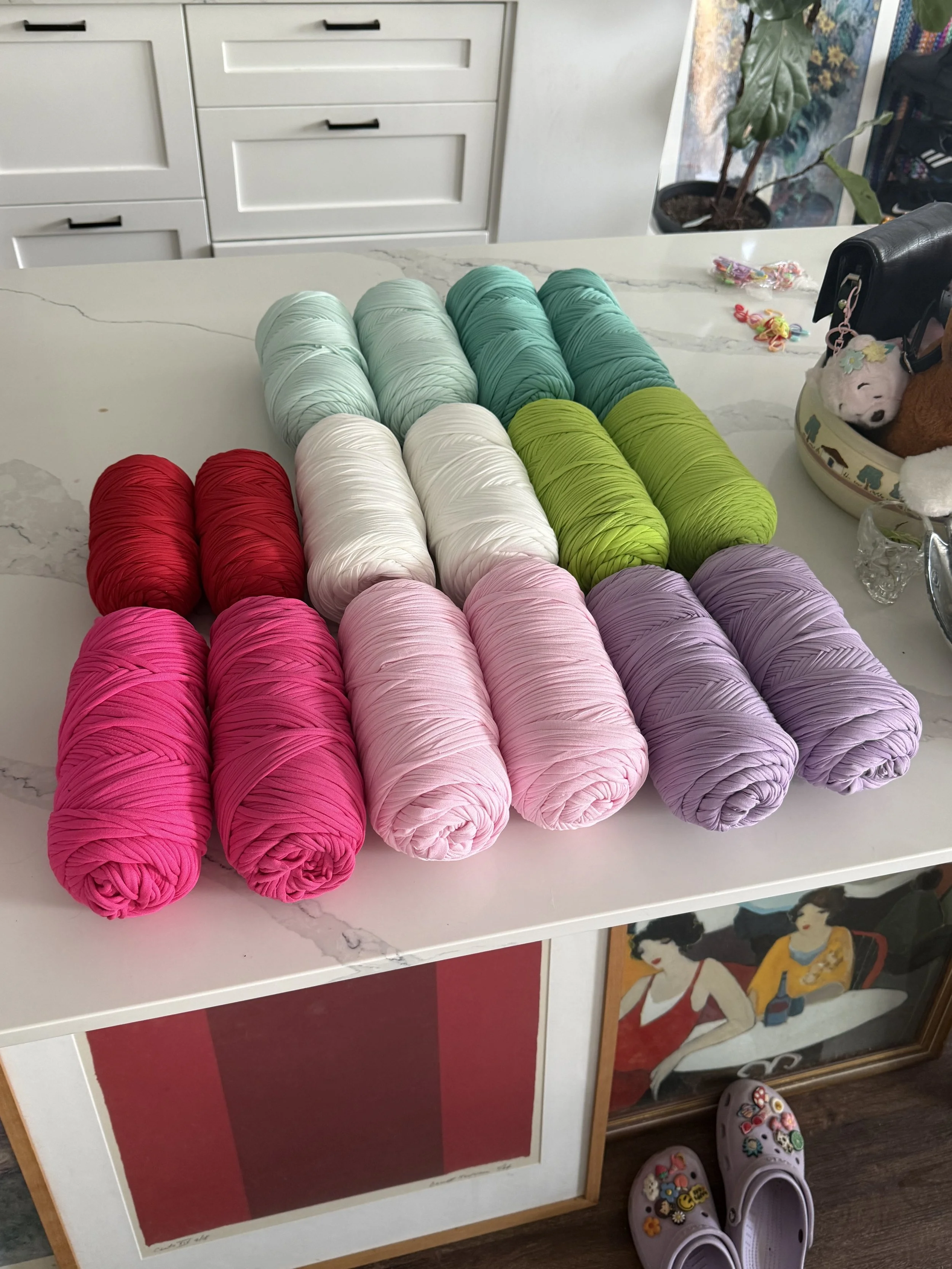 Several spools of yarn in various shades of pink, white, green, and blue arranged on a white table. A pair of purple Crocs decorated with various colorful pins and charms are on the floor beneath the table. In the background, there's a white cabinet, a potted plant, and some small colorful hair ties or rubber bands on the counter.