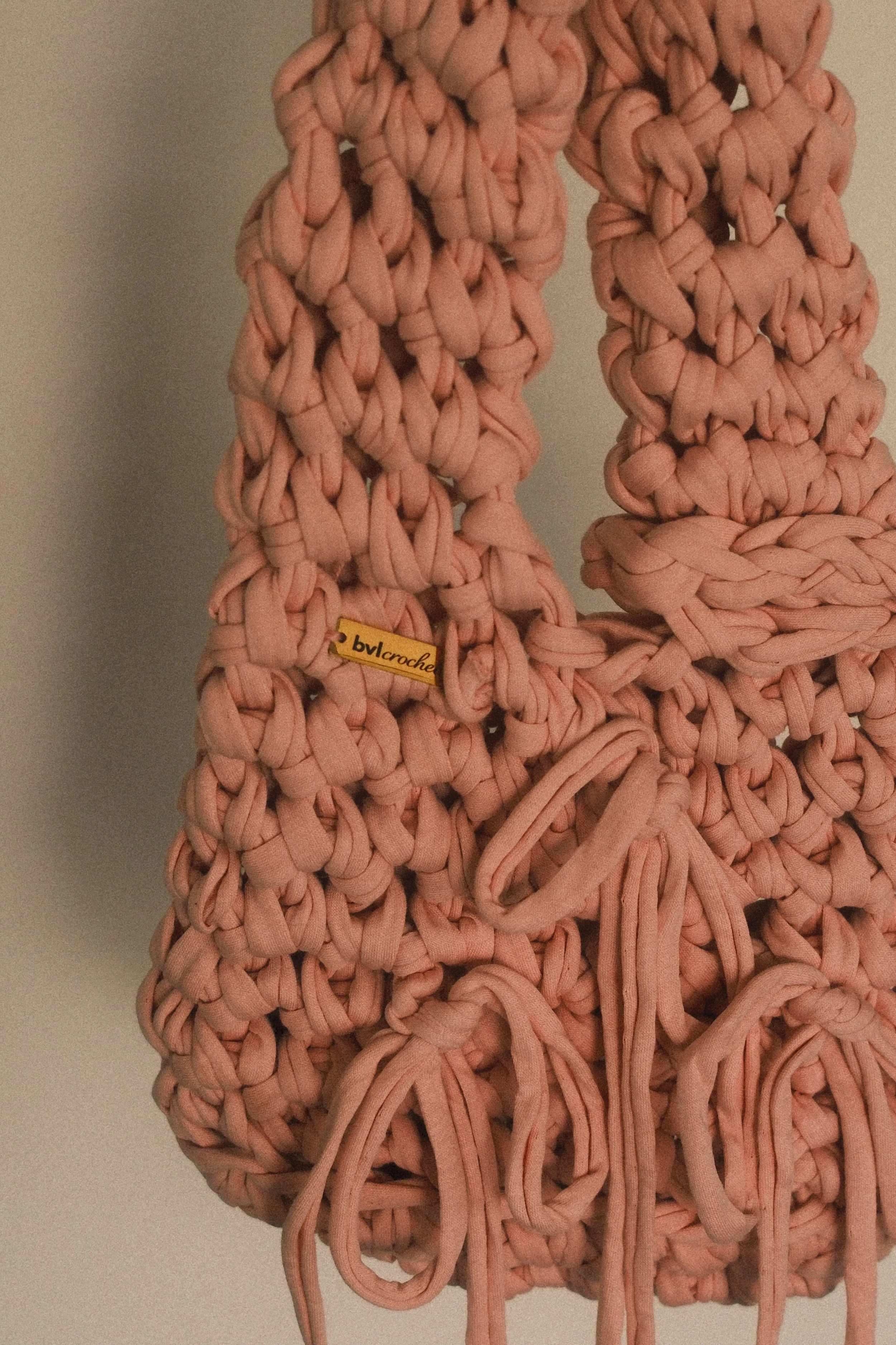 Close-up of a large handmade crochet item in pink yarn, showing intricate knot and weave patterns, with a small yellow tag labeled 'bvlcroche' attached.