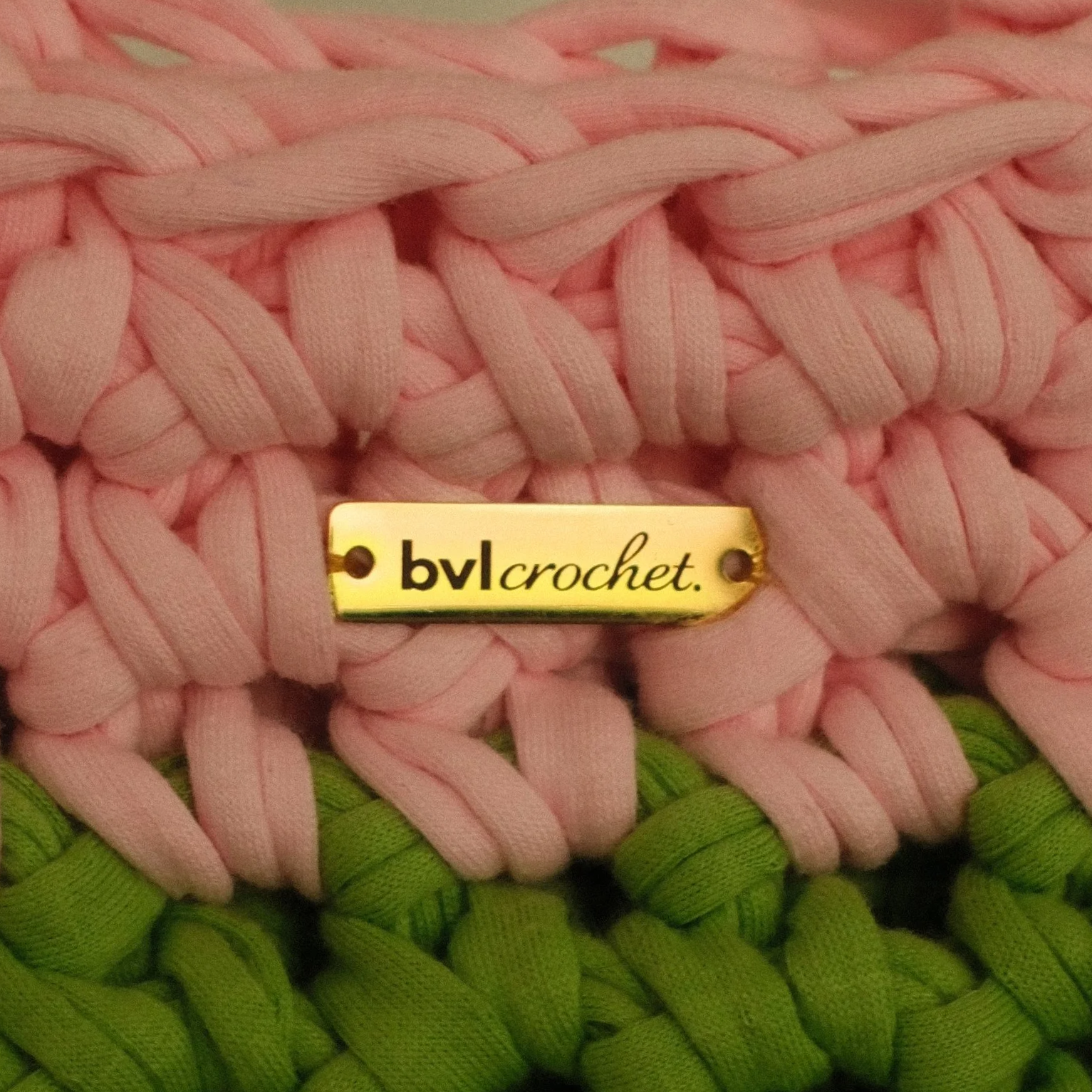 Close-up of a pink and green woven crochet piece with a gold tag reading 'bvl crochet'.