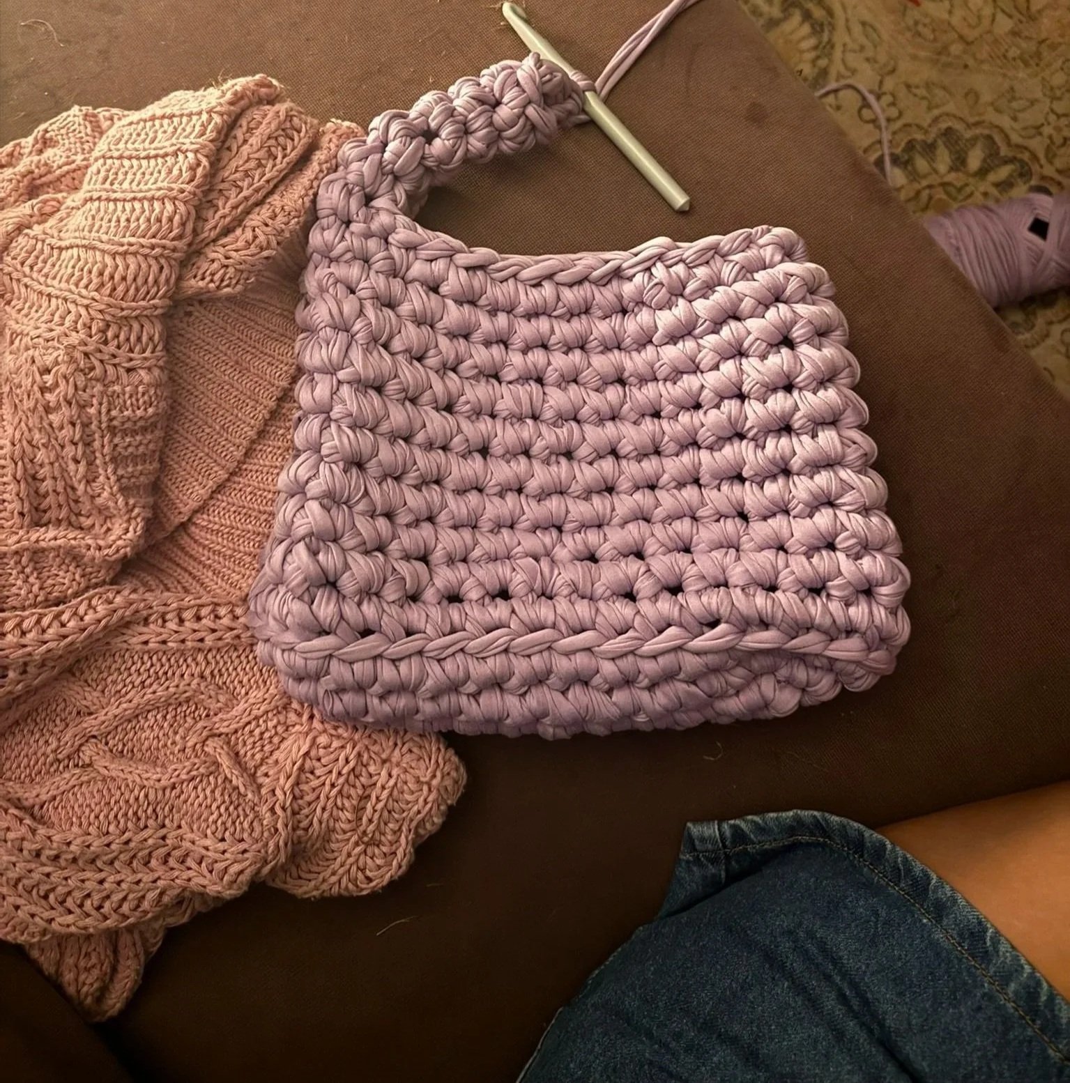 A pink knitted blanket, a light purple woven bag, and a pink knitted sock on a brown couch with a patterned rug in the background. A person's leg wearing denim shorts is visible in the bottom right corner.