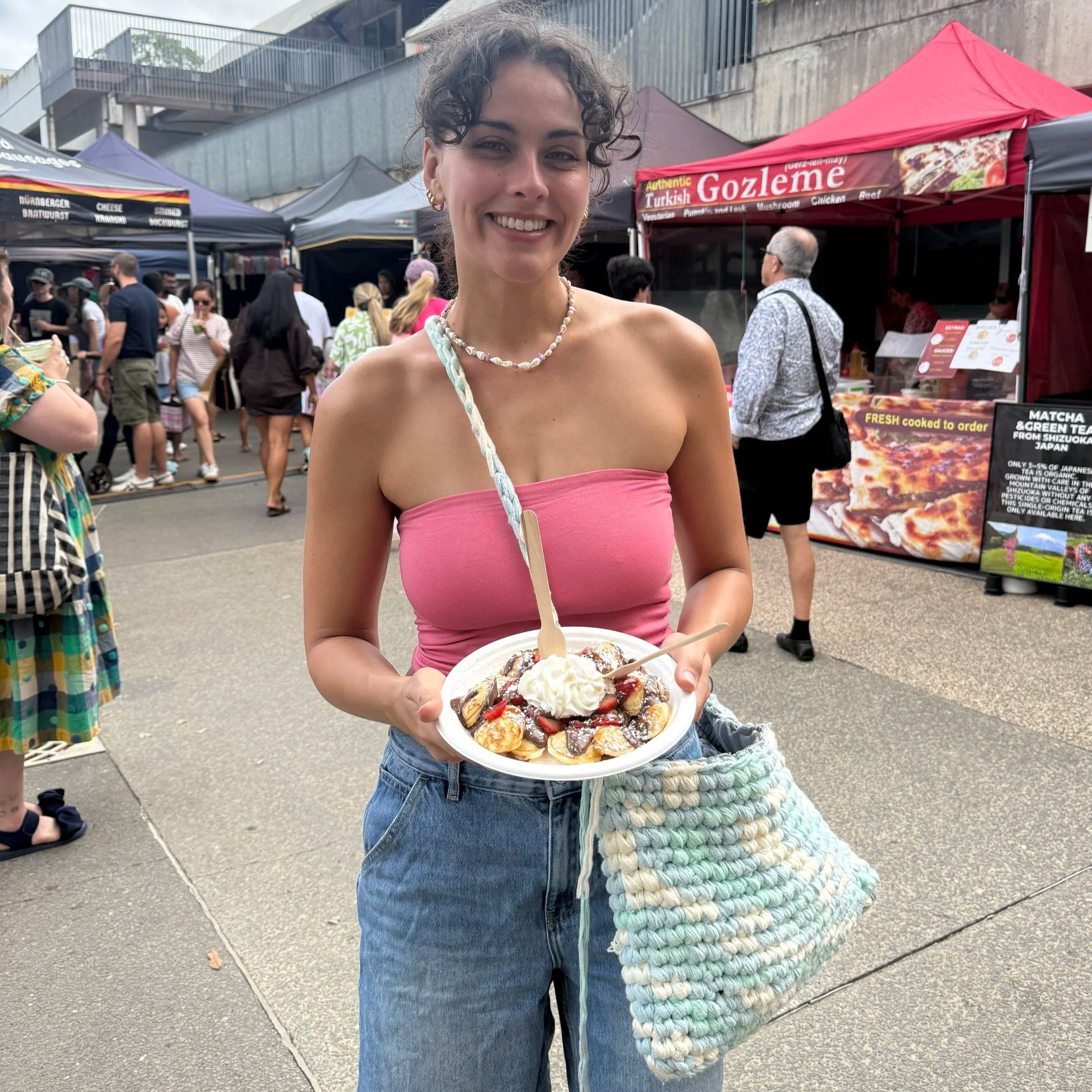 A young woman smiling and holding a plate of dessert, standing outdoors at a market with food tents and other people in the background.