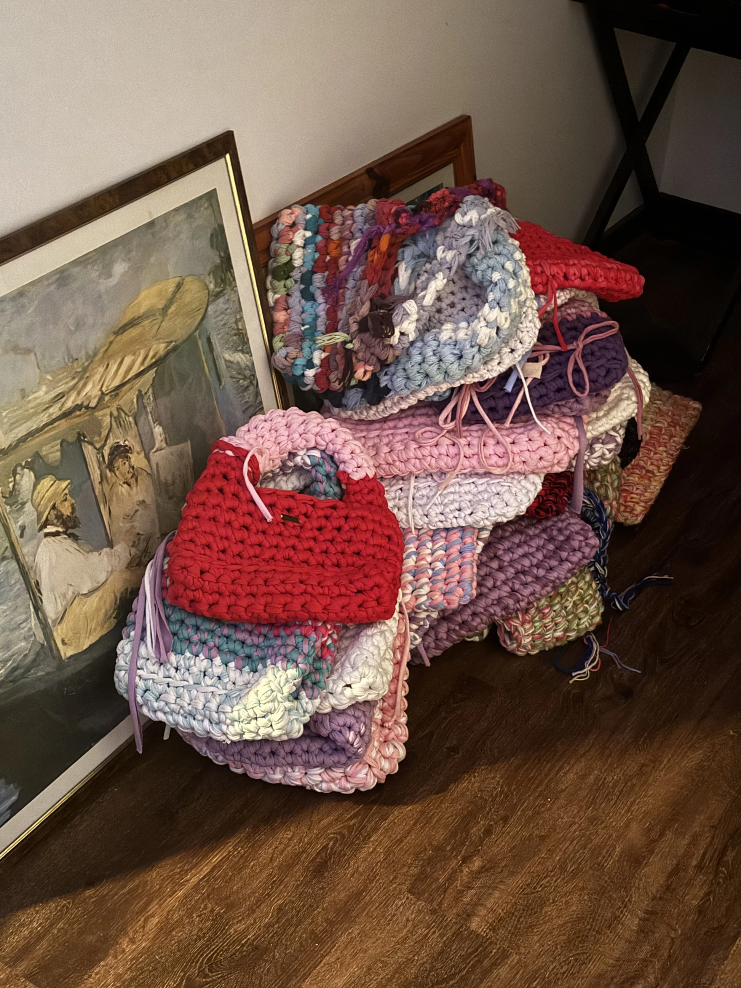 A stack of colorful crocheted blankets and scarves placed on a table next to a wooden framed painting leaning against the wall.