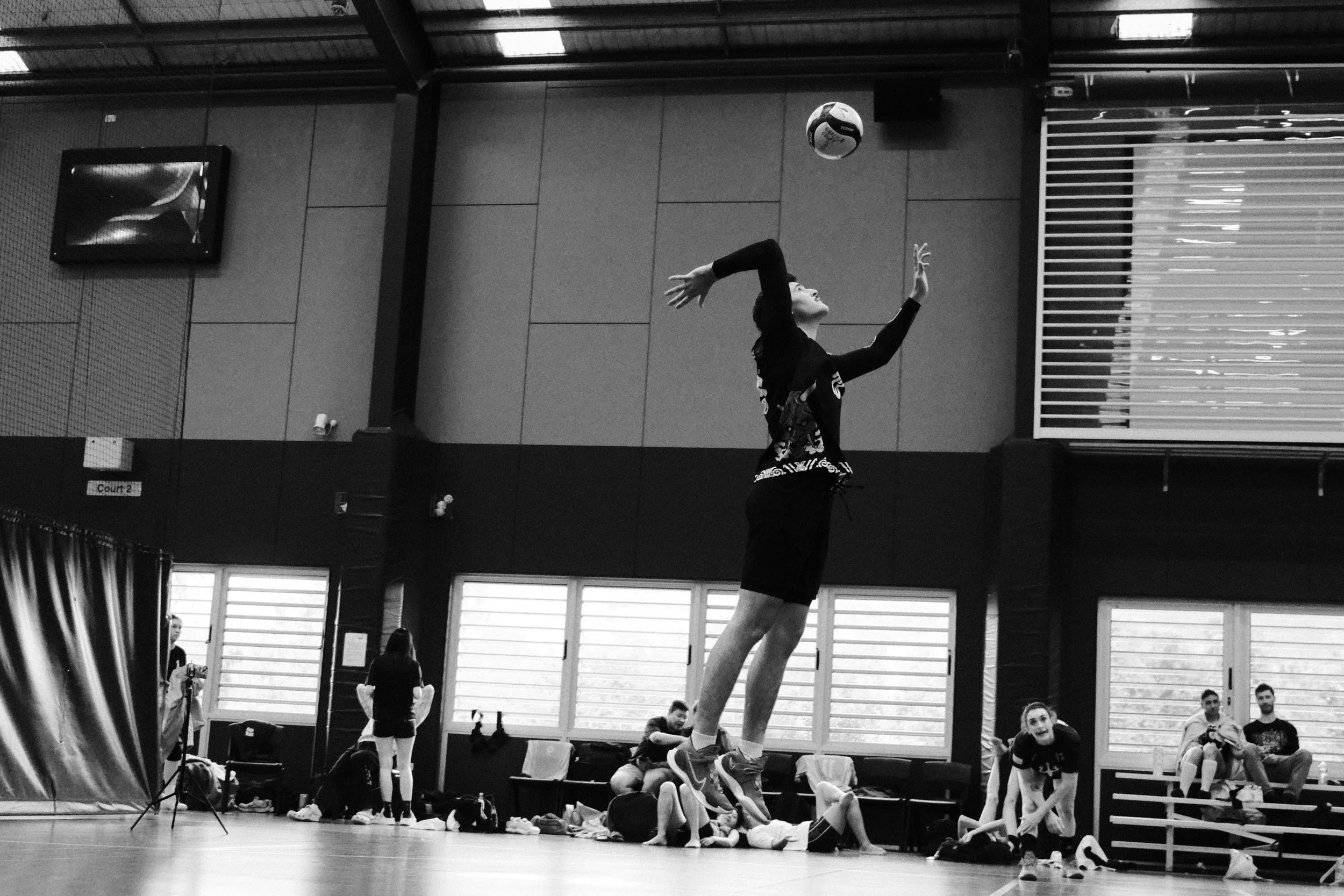 A woman jumps to hit a volleyball in an indoor gym, with several other people sitting and standing around watching.