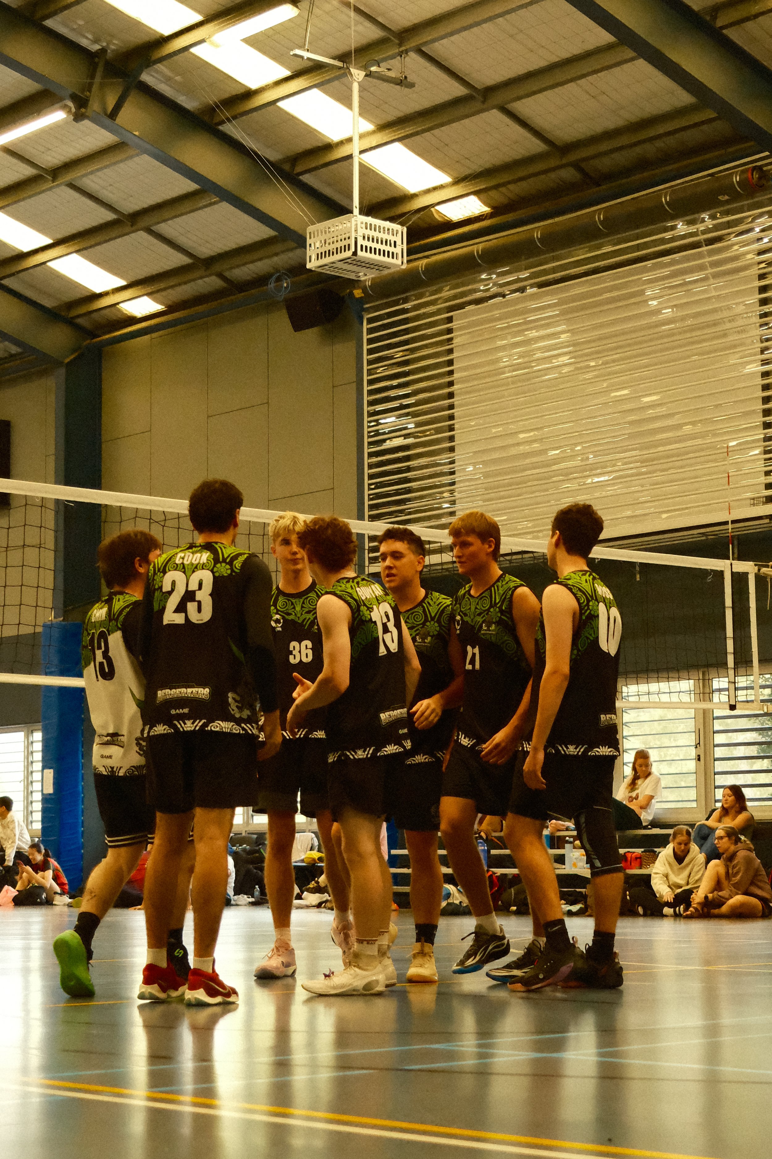 A group of volleyball players in black and green uniforms gathered in a circle on an indoor court, possibly during a timeout or team discussion. The gym has high ceilings with lighting and some spectators sitting in the background.