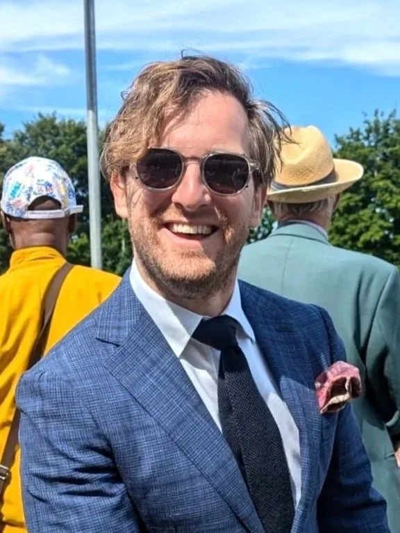 A smiling man in sunglasses, a blue suit, white shirt, and dark tie, standing outdoors during sunny weather, with two other people in the background.