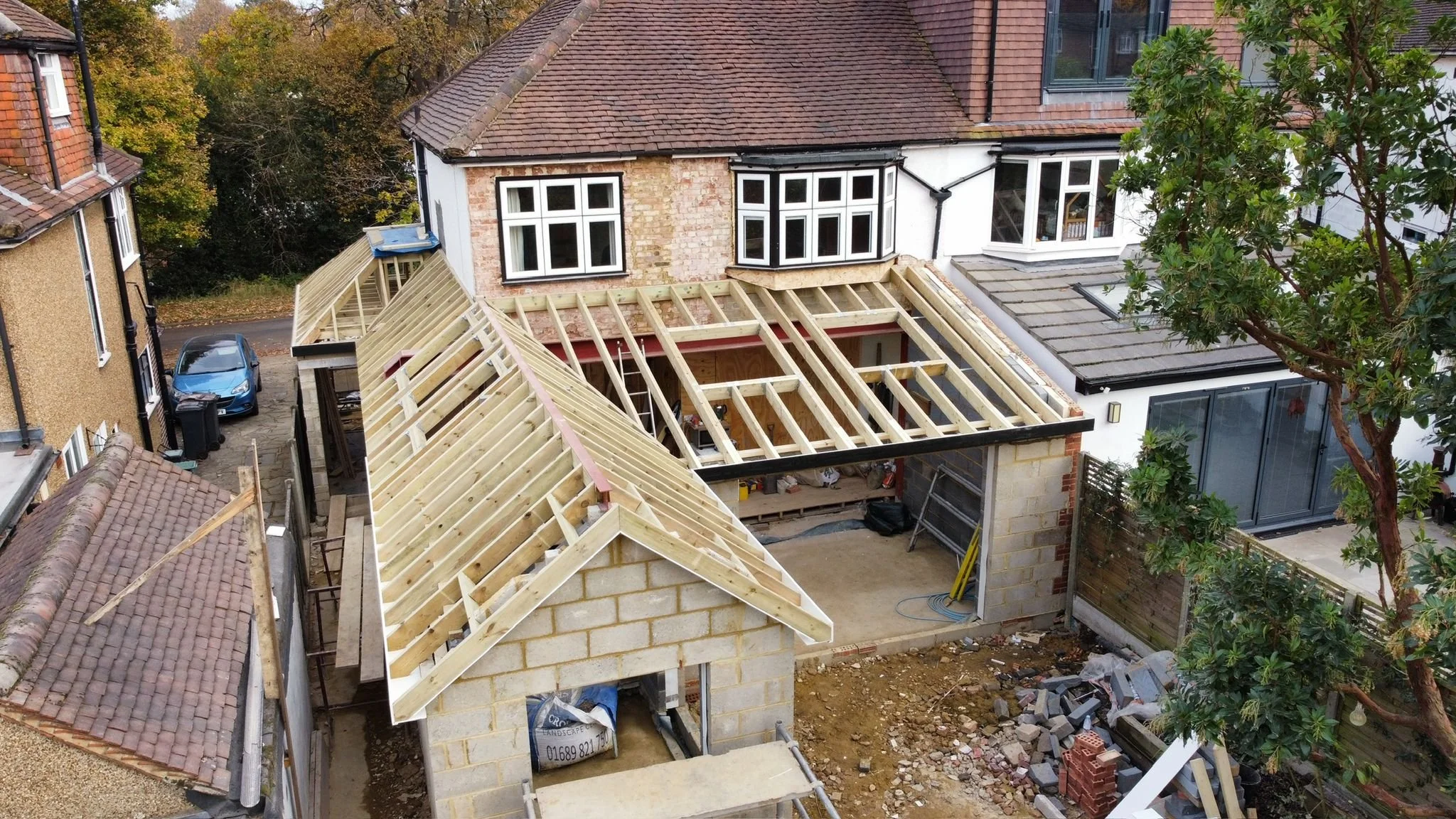 Construction site of an extension to a residential building with an unfinished roof, wooden framing, and surrounding houses and trees.