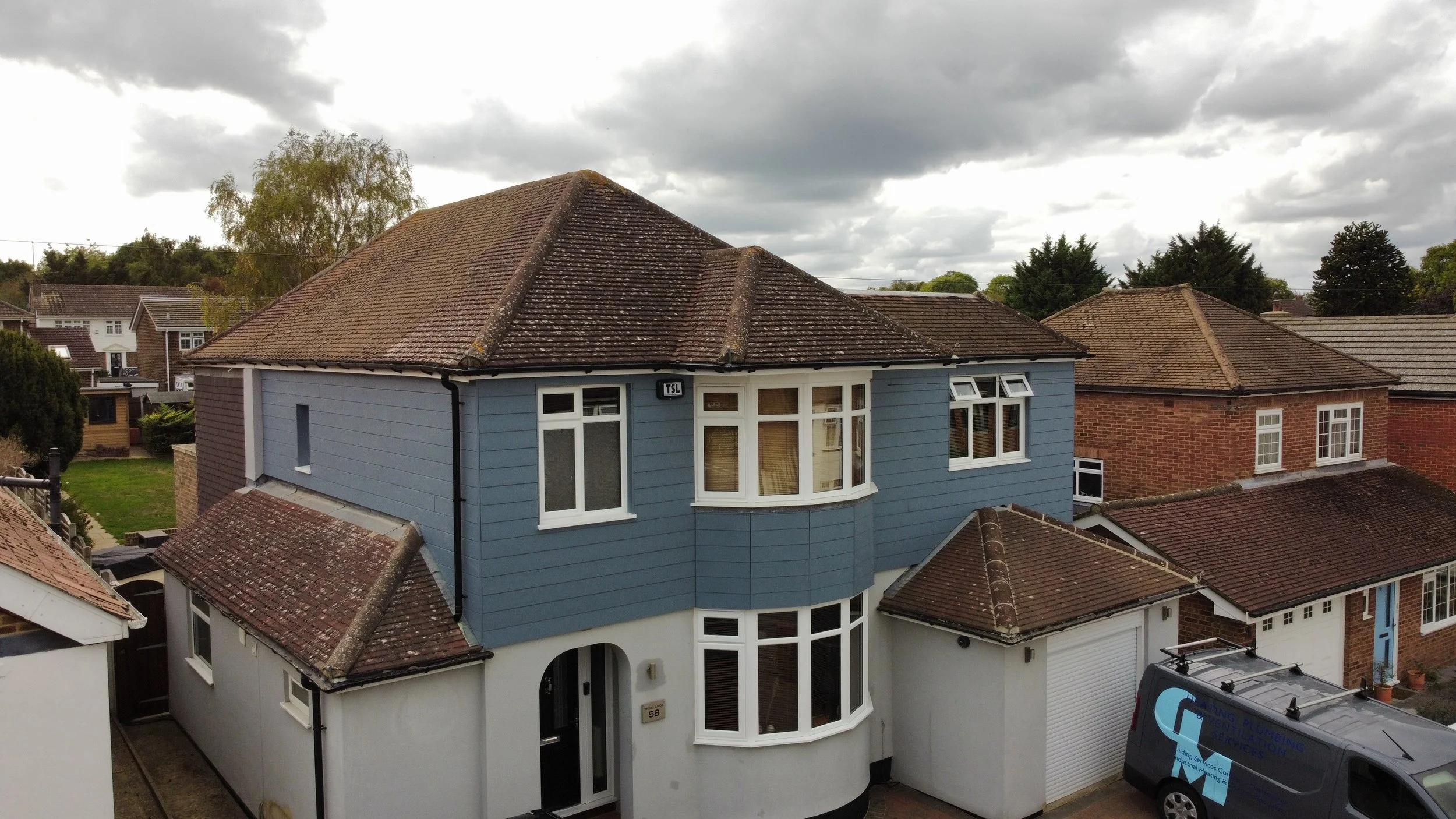 A two-story house with a blue exterior, white trim, and a curved bay window on the upper floor. The house has a brown tiled roof and is situated in a neighborhood with other houses and trees visible in the background under a cloudy sky.