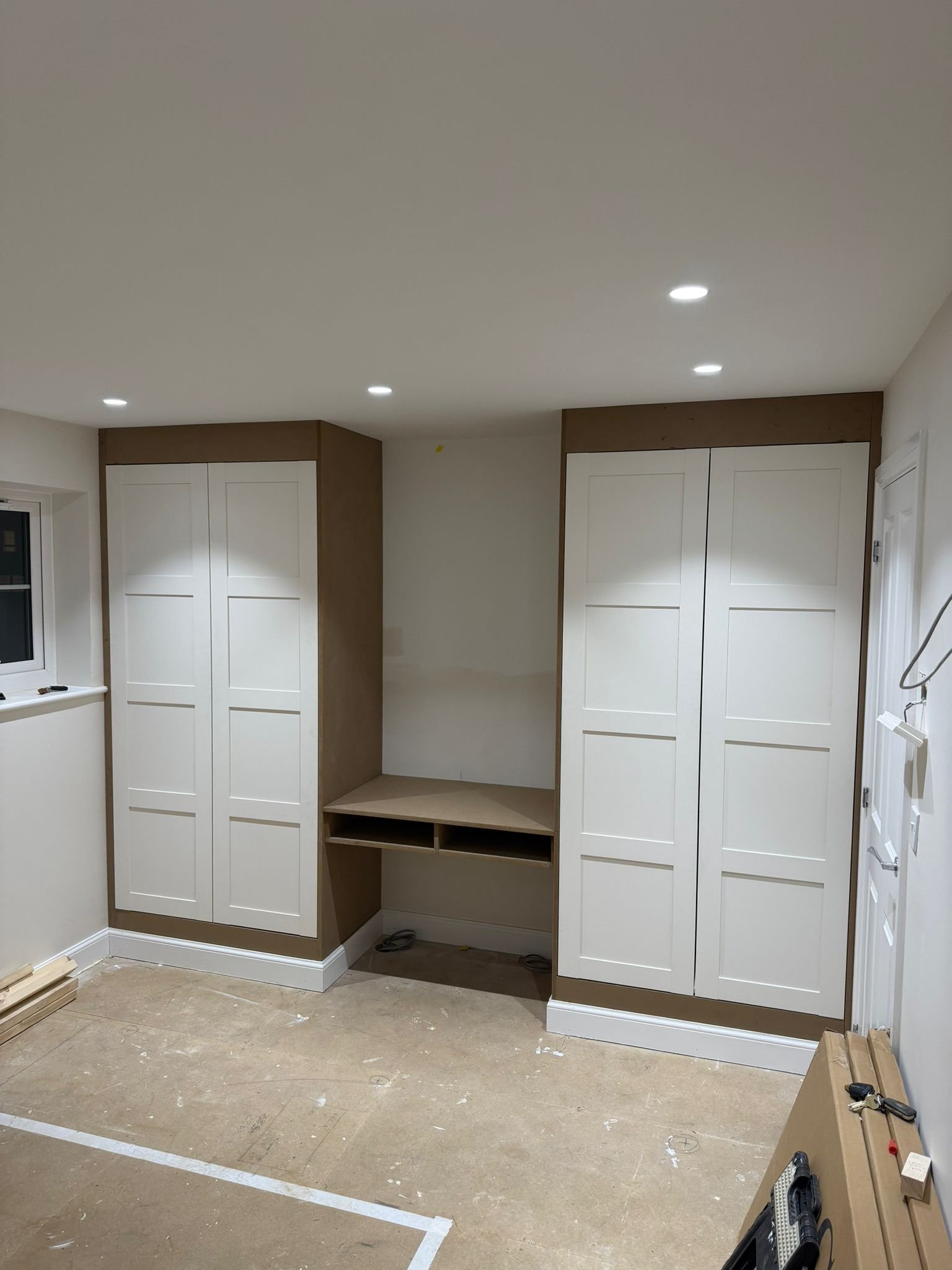 A partially constructed room with white cabinets, a window, and a rough floor. Construction tools and supplies are visible.