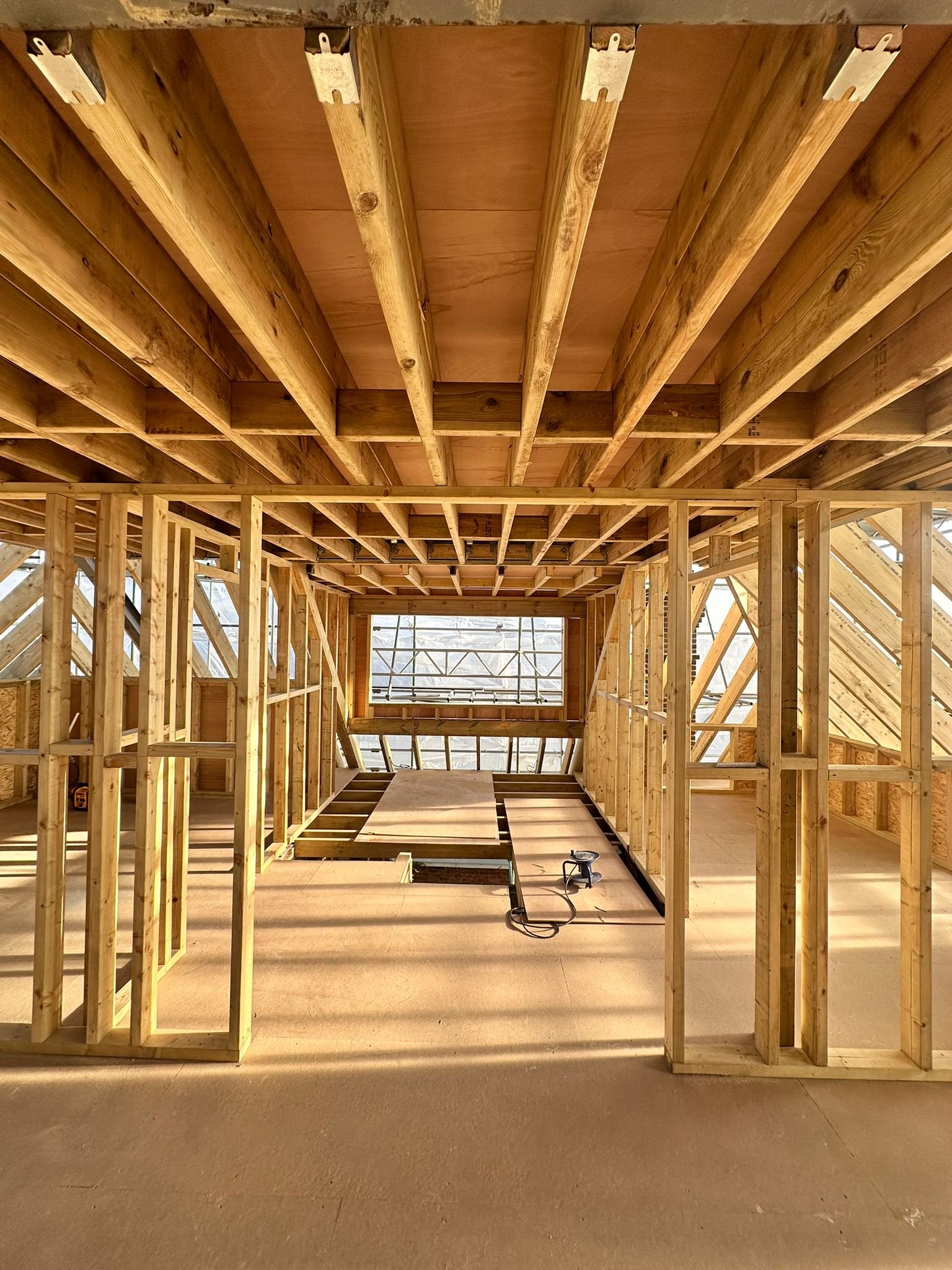 The interior of a house under construction, showing wooden framing, a partially finished ceiling, and a window at the back.