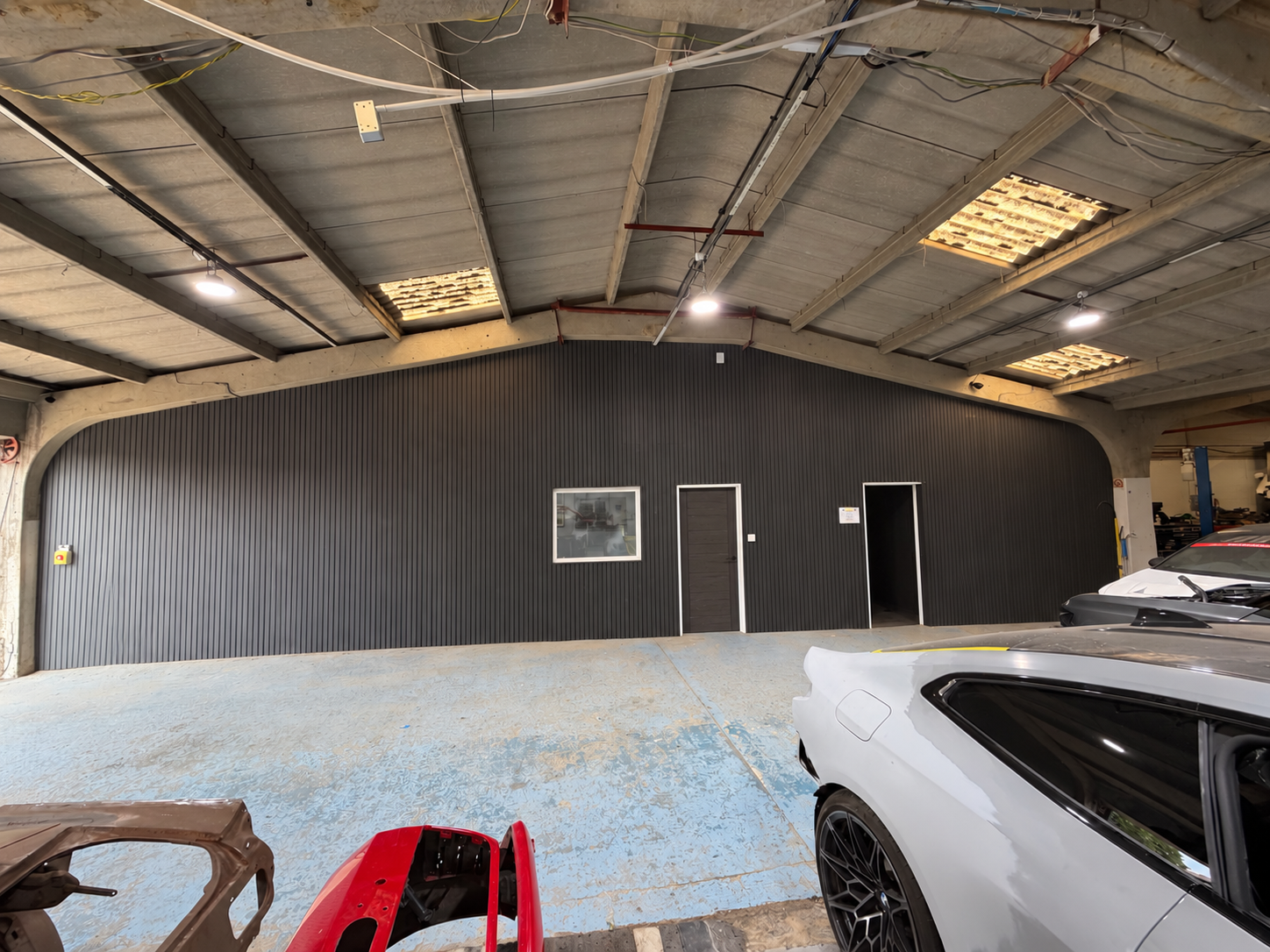 An indoor parking area with unfinished ceiling, black corrugated wall, a small window, a door, and a doorway leading to another part of the garage, with several cars parked.