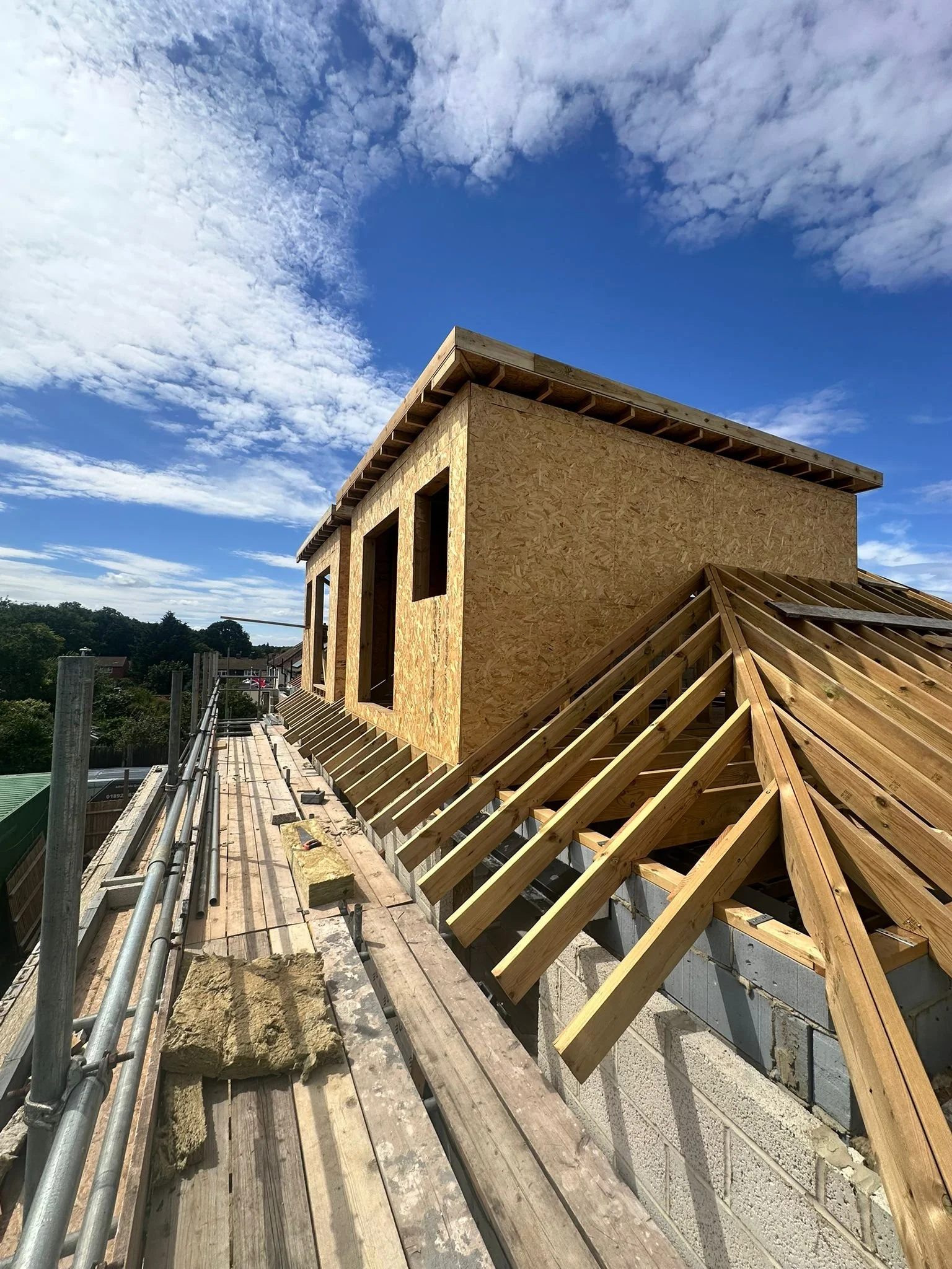 Construction site with a house under construction made of plywood and wooden beams, scaffolding, and a partly cloudy blue sky.