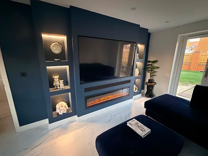 Living room with dark blue accent wall, mounted flat-screen TV, built-in shelves with decorative items, electric fireplace, and a sliding glass door leading outside.