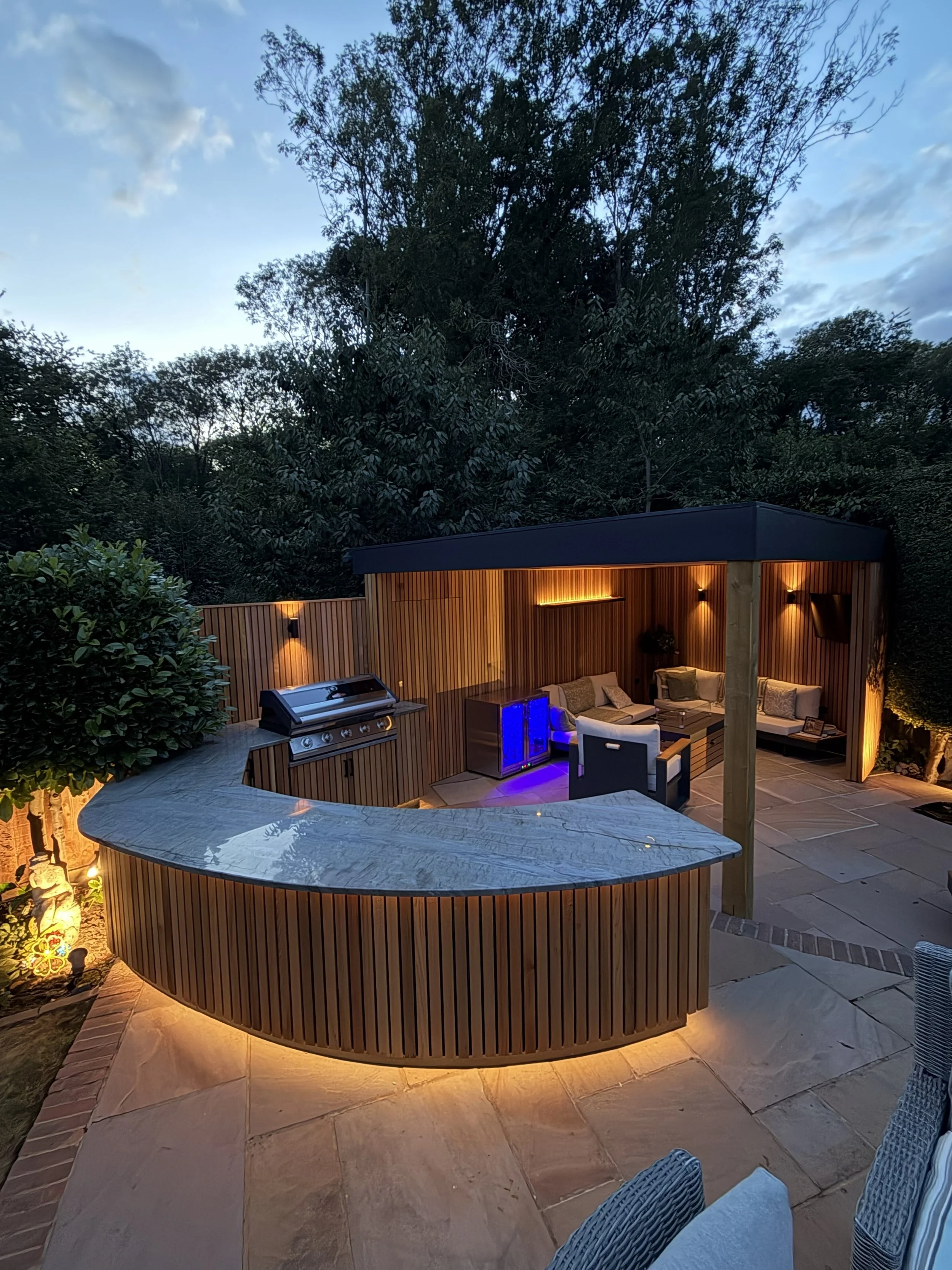 An outdoor patio area with a curved marble counter, a grill, a wooden screened seating nook with outdoor furniture, blue accent lighting, surrounded by lush greenery and trees, during dusk.
