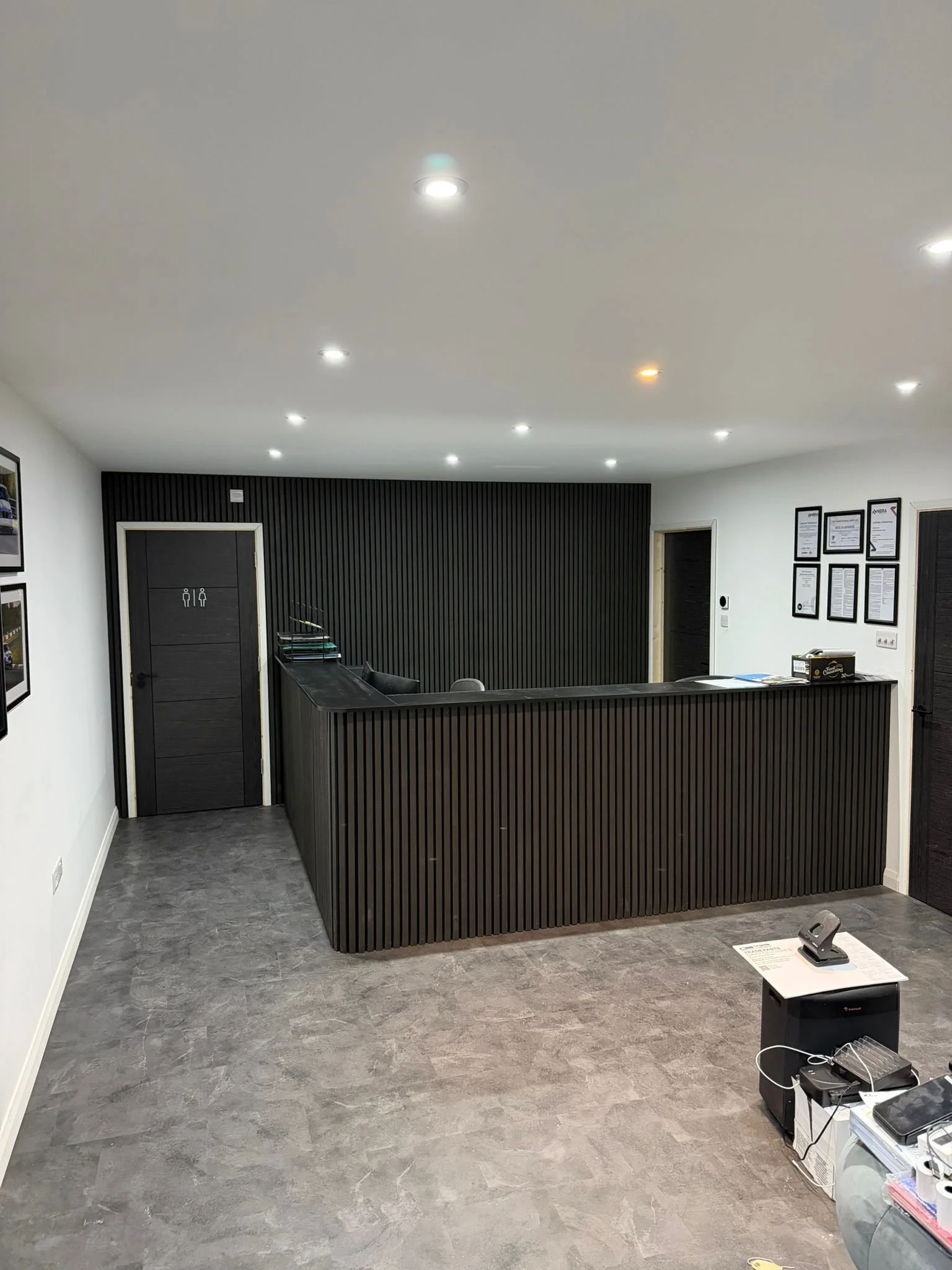 Modern reception area with black reception desk, dark wall panels, framed papers on white walls, gray flooring, and a small table with office supplies and electronic devices.