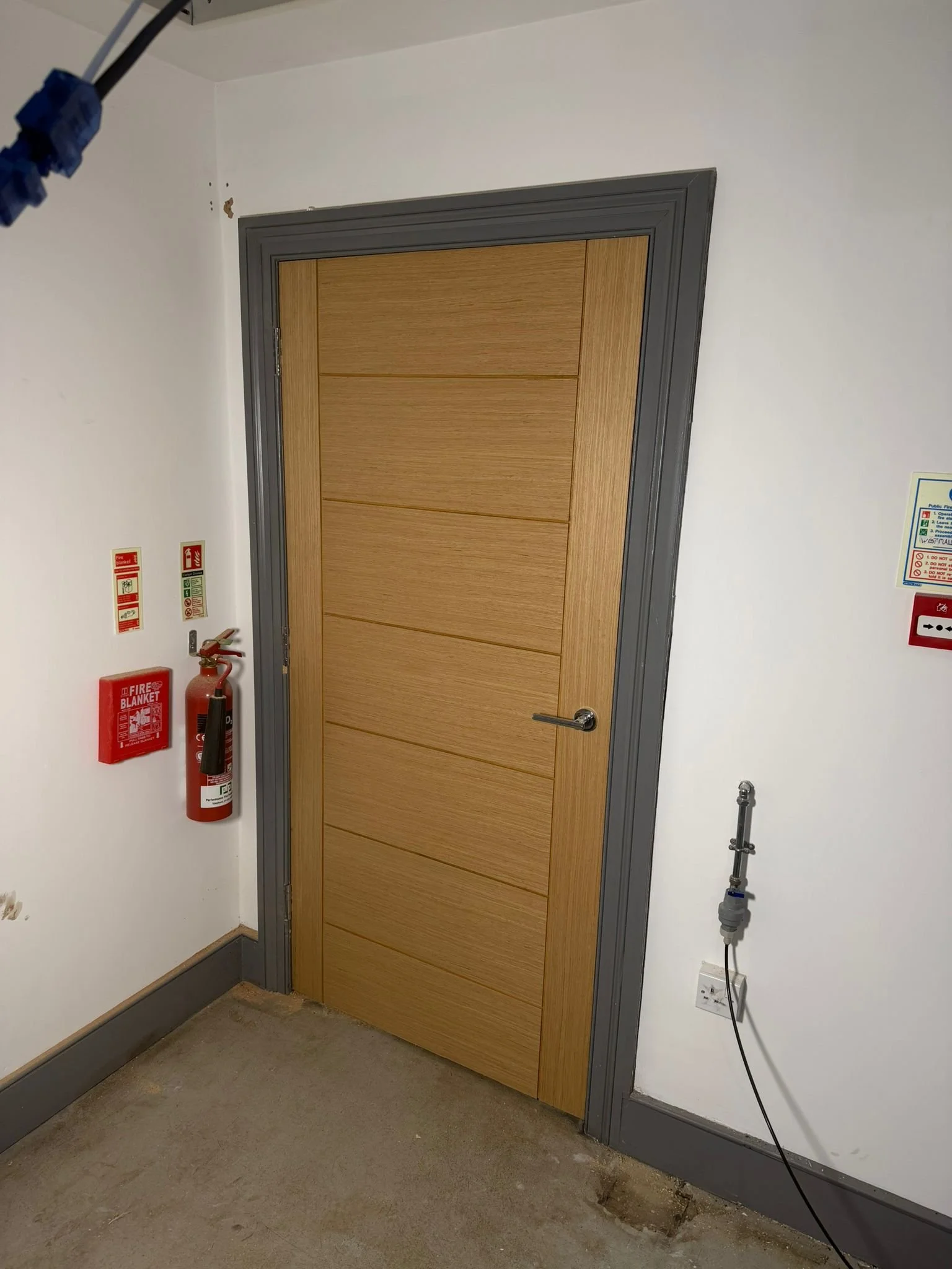 Interior of a building with a closed wooden door and gray frame. Fire safety equipment including a fire blanket and extinguisher mounted on the wall to the left of the door. Safety and fire warning signs on the wall near the equipment. An electrical outlet and visible wiring at the bottom right.
