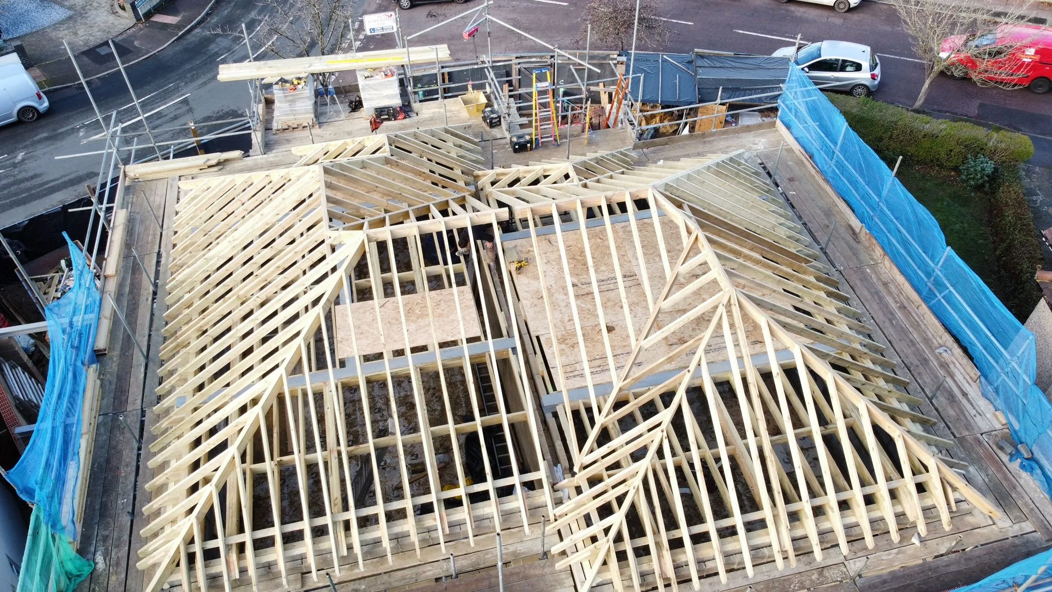 Construction site view from above showing the wooden framework of a building with a gabled roof, surrounded by scaffolding and construction tools, with parked cars and street in the background.