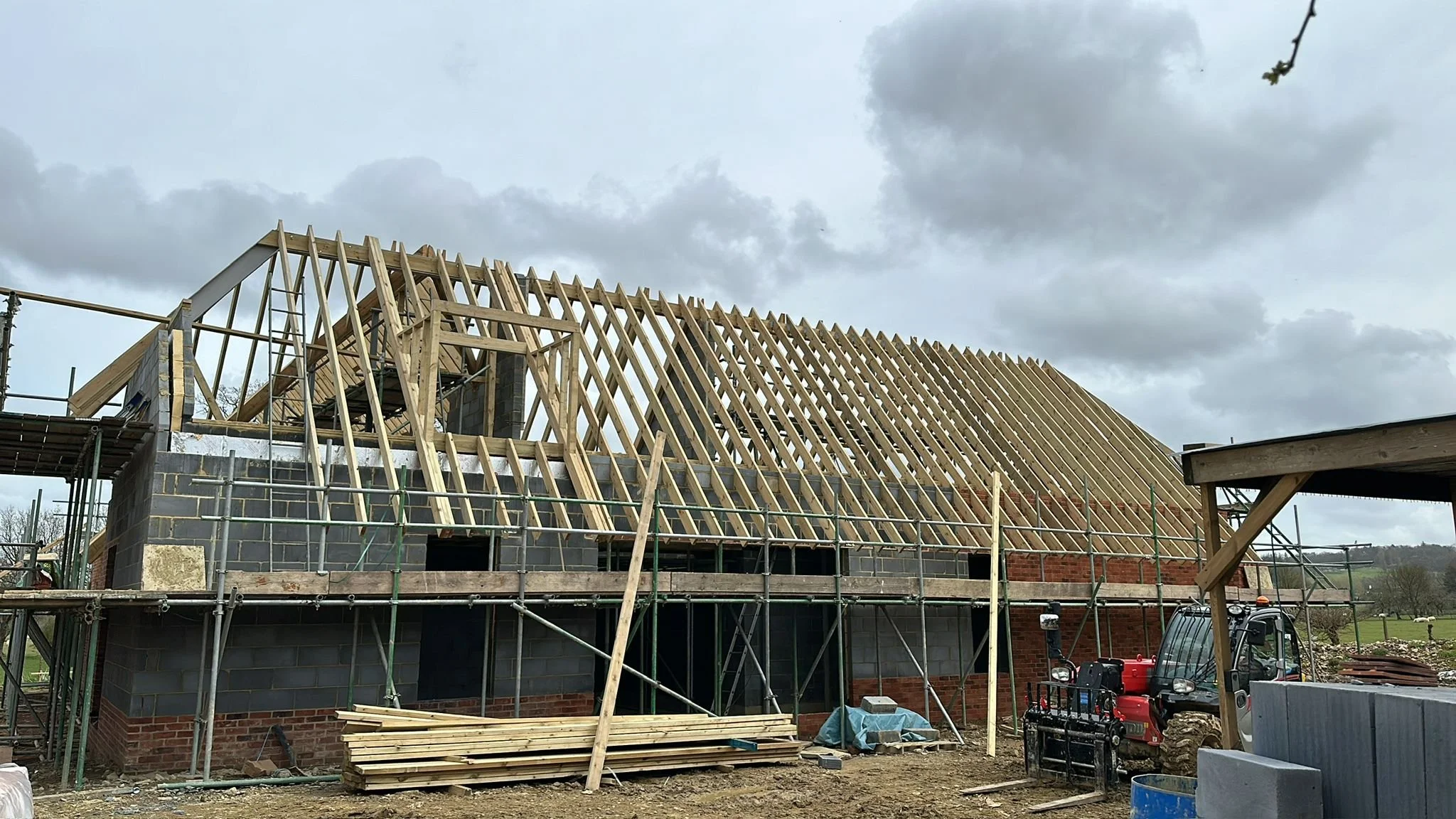 Construction site with a house in the process of being built. Wooden roof framework is visible, with some scaffolding around the building. The sky is cloudy.