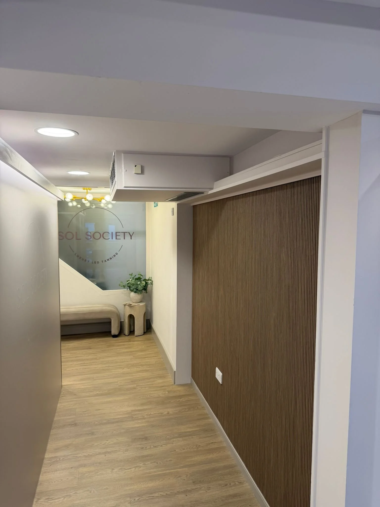 Interior of a modern office or clinic with wood flooring, a wood-paneled wall, a small seating area with beige chairs and a small side table with a potted plant, and a door with the logo 'SOL Society' and the words 'Luxury LED Tanning' on a frosted glass panel.
