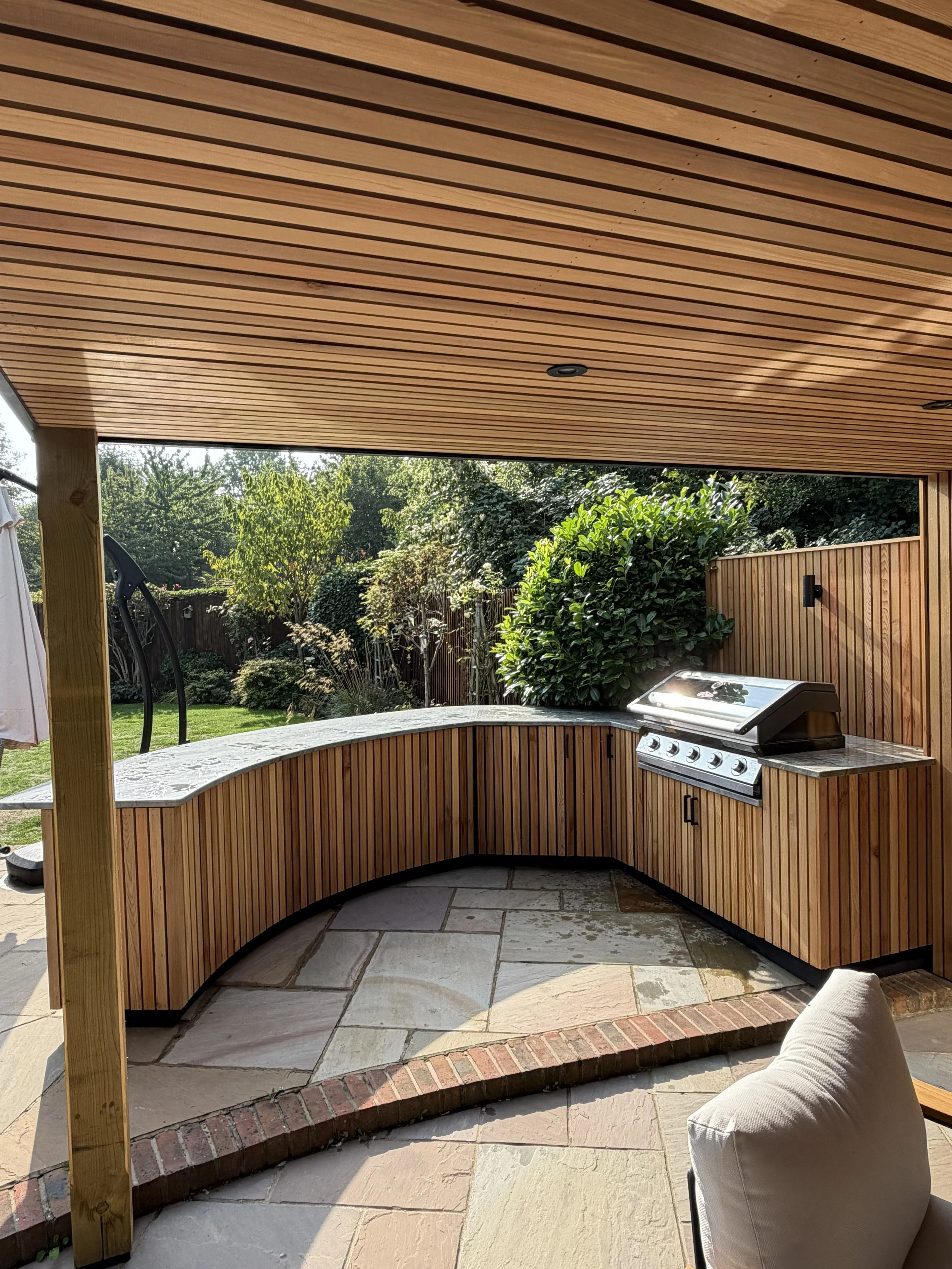 Outdoor kitchen with a built-in grill, curved wooden cabinets, and a stone patio, surrounded by greenery and a wooden privacy fence.