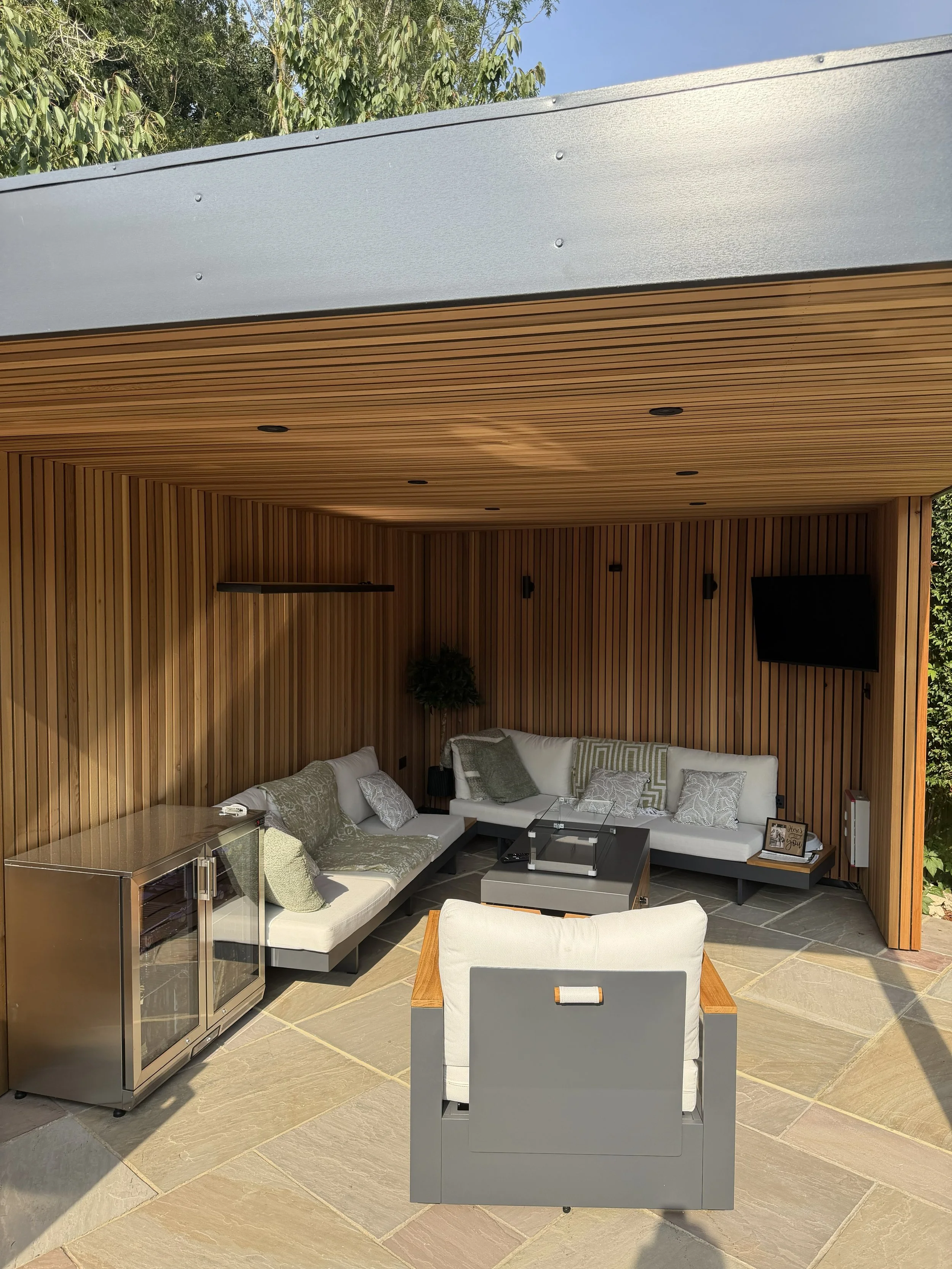 A modern outdoor living space with a wooden-paneled ceiling and walls, a white sectional sofa with pillows, a small glass-top coffee table, a stainless steel mini fridge, a white armchair, a small side table, a mounted flat-screen TV, and outdoor lighting.