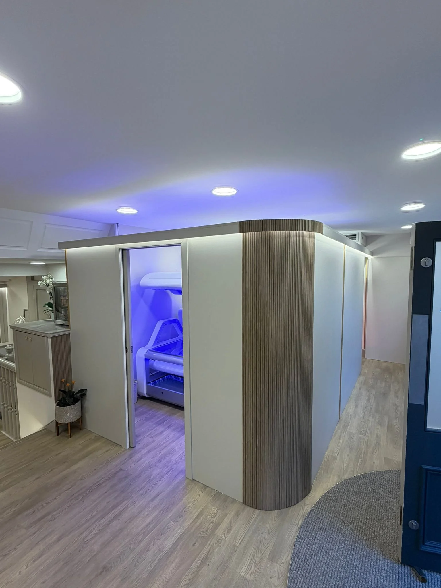 A modern indoor space with a wooden-paneled curved wall and a white enclosed booth with blue lighting. The ceiling has recessed lights, and the floor is light-colored wood. There's a small table with a plant in a pot in the corner.