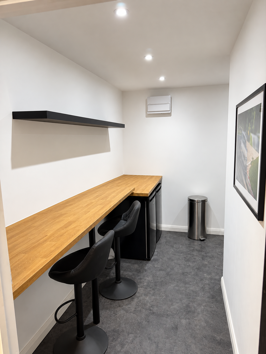 Small minimalist kitchenette with a wooden countertop, two black bar stools, a black mini fridge, a stainless steel trash can, a wall-mounted black shelf, and white walls with a framed picture.