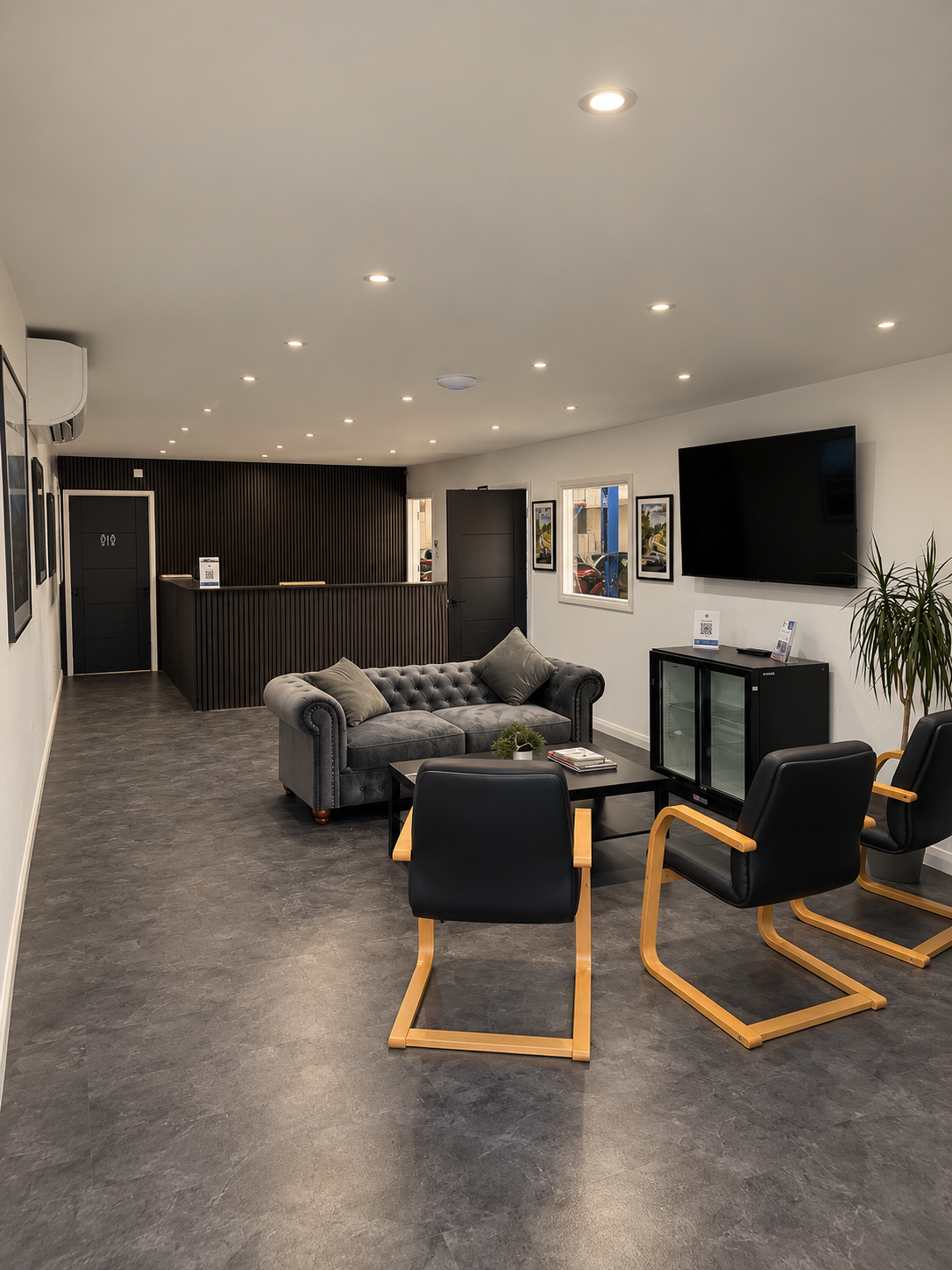 Modern hotel lobby with a gray velvet sofa, black lounge chairs with wooden arms around a black coffee table, a flat-screen TV on the wall, and a reception desk in the background.