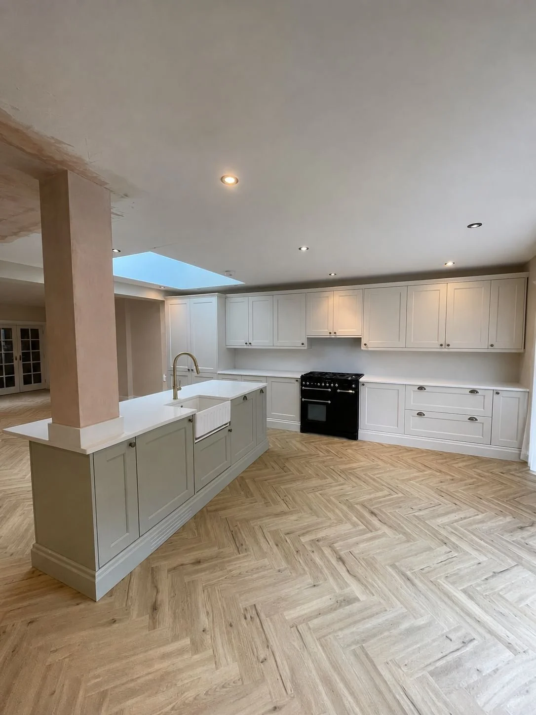 Modern kitchen with white cabinets, a black stove, a kitchen island with a sink and gold faucet, light wood herringbone flooring, and a skylight in the ceiling.