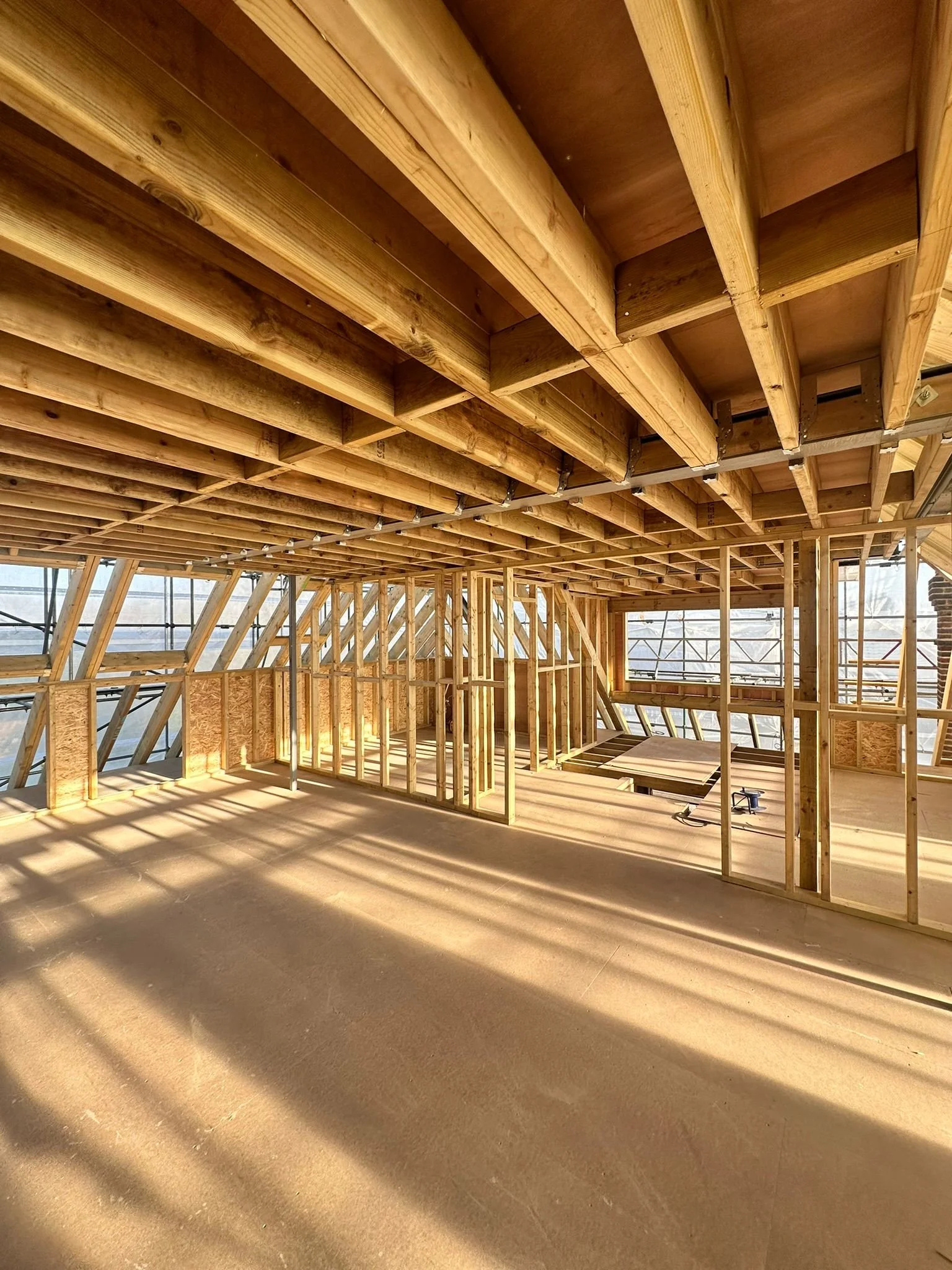Interior view of a building under construction with exposed wooden framework and beams, and sunlight casting shadows on the unfinished floor.
