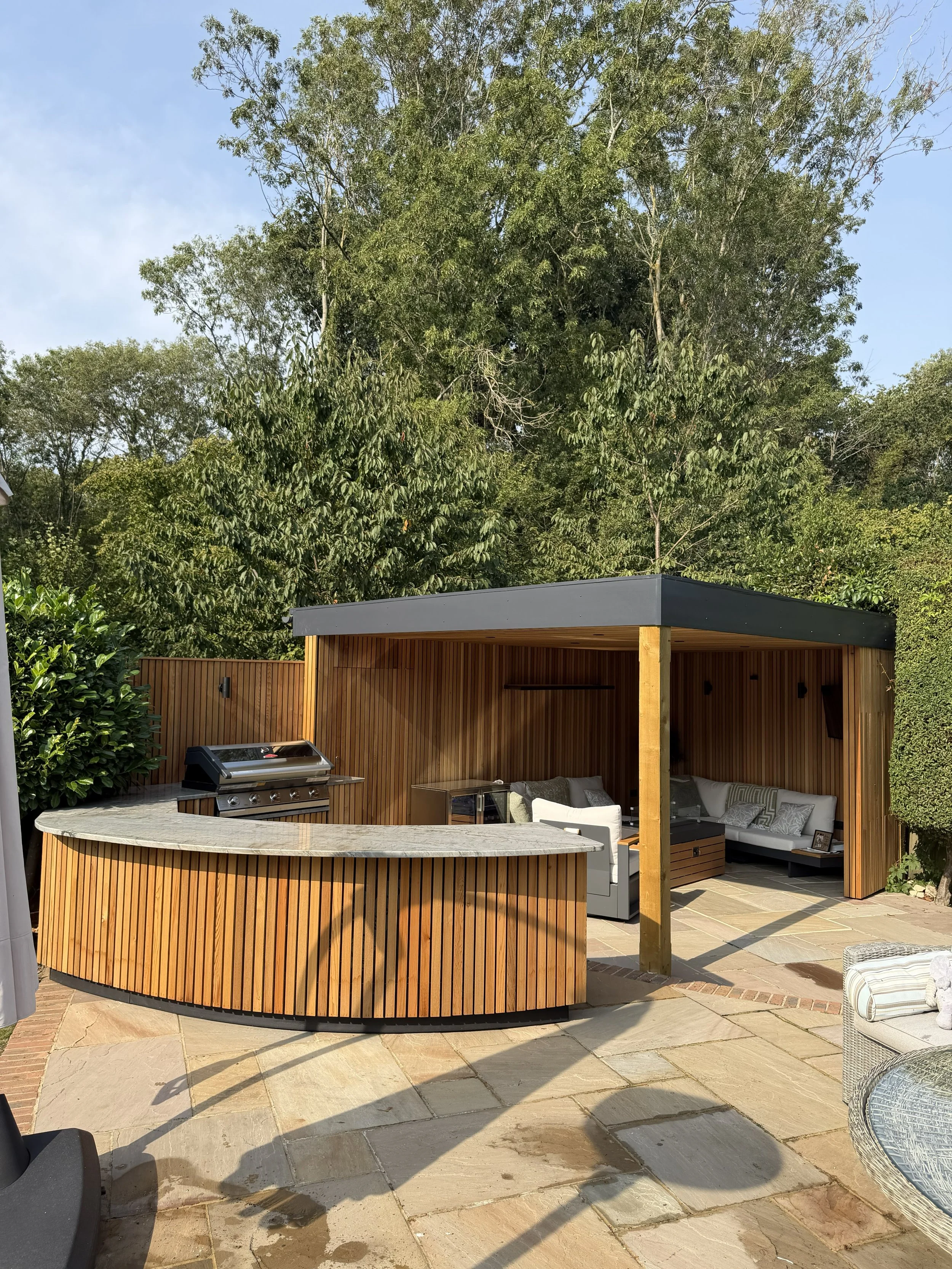 Outdoor patio area with a built-in wooden counter, a grill, and a covered seating area with couches and pillows, surrounded by trees and greenery.