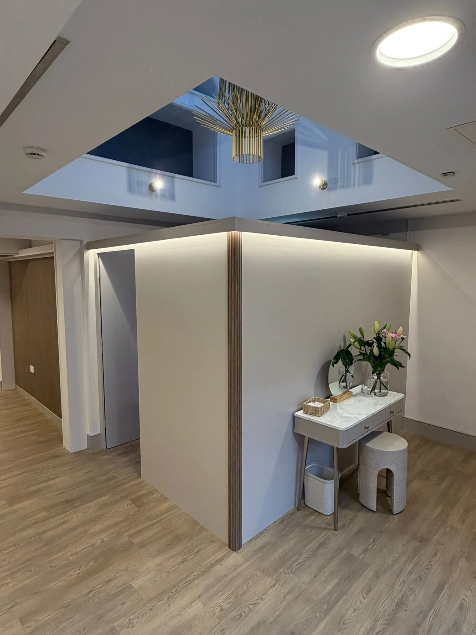 Interior of a modern room with a small white desk, a gray stool, and a potted plant with pink flowers on the desk. There is a partial wall dividing the space, and a decorative hanging light fixture above. The ceiling has a cut-out section with ceilin
