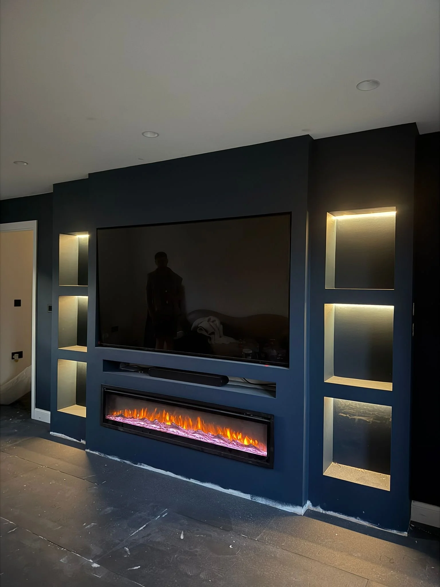 Modern indoor entertainment wall with a large flat-screen TV, built-in electric fireplace, and illuminated shelves on either side, in a room under construction or renovation.