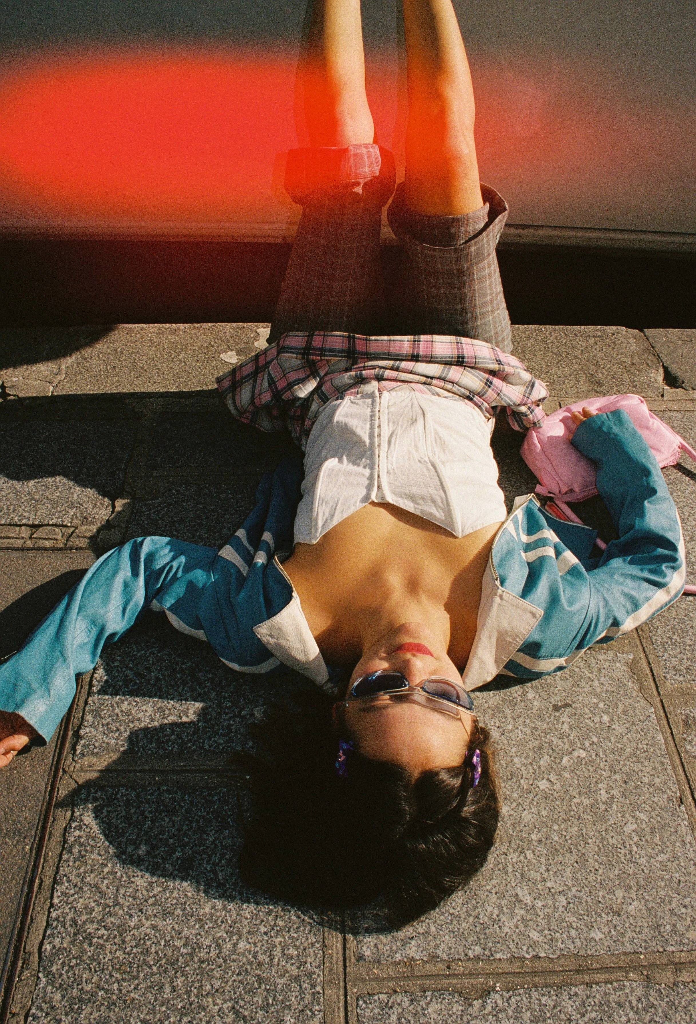 A person lying on a sidewalk wearing sunglasses, a partially unbuttoned jacket, and a white shirt revealing their chest. They have dark hair and are wearing a headband with a bow. Their arms are extended outward, and they have a pink bag and a teal item nearby.