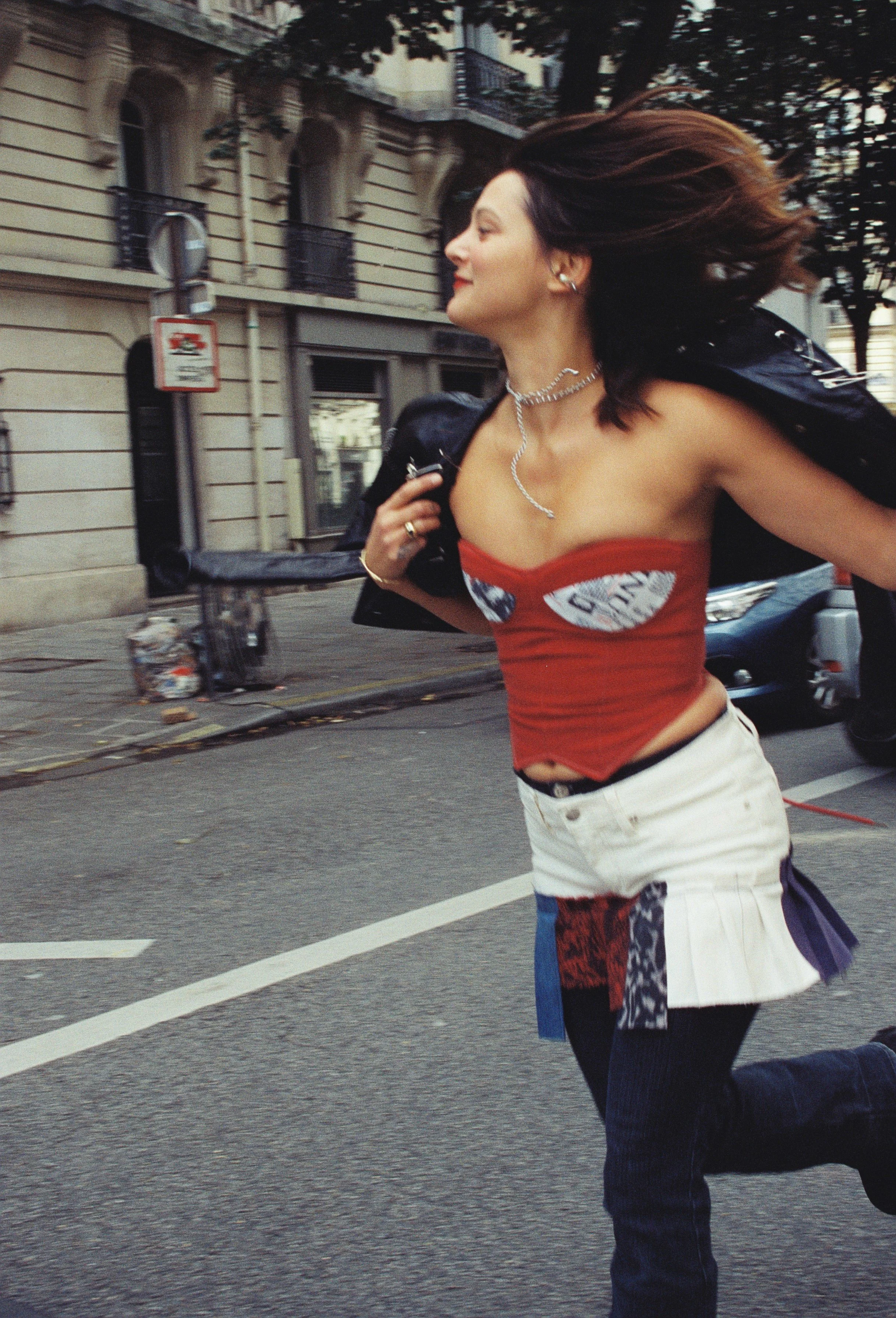 A young woman with dark brown hair flowing behind her is running on a city street. She is wearing a sleeveless red top with graphic patches, a silver necklace, and earrings, and has a black jacket draped over her shoulders. The background shows a city building with balconies, trees, a street sign, and a bicycle.