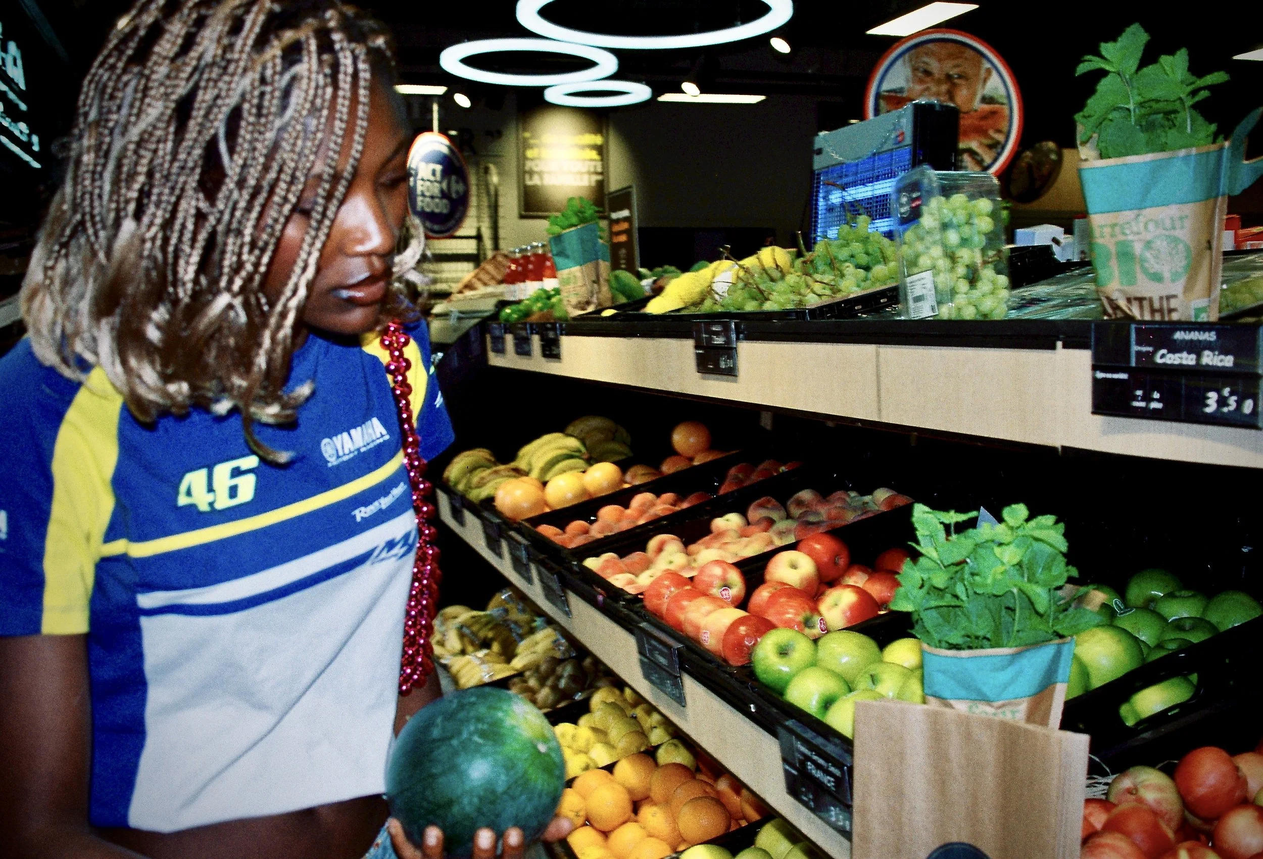 A woman with braided hair wearing a blue and yellow sports jersey shopping in a produce section, holding a watermelon. The shelves behind her are filled with various fruits such as apples, grapes, and bananas, in a grocery store.