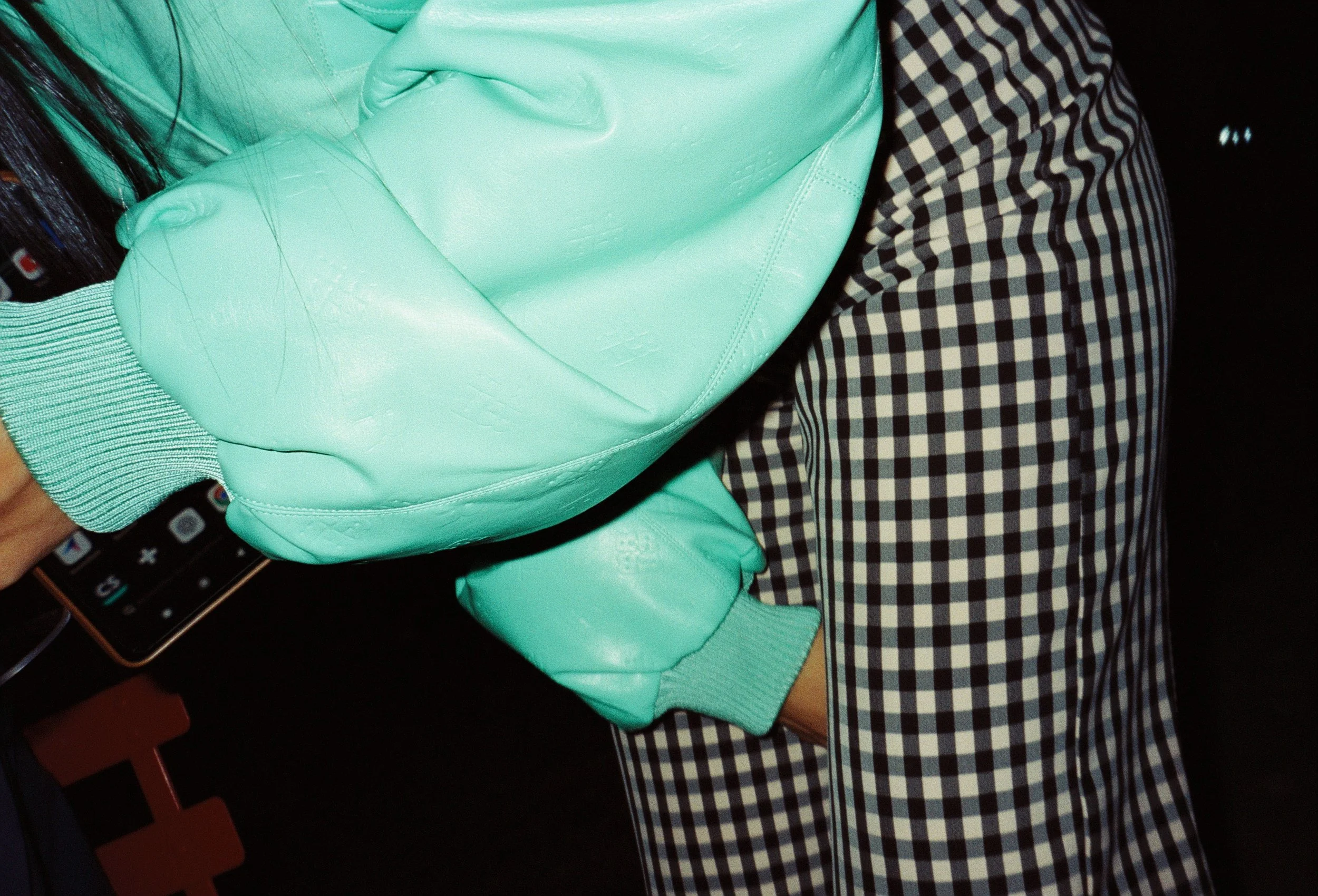 A person wearing a mint green jacket and black-and-white checkered pants is holding a black smartphone with app icons visible on the screen. The person's long dark hair is partially visible.