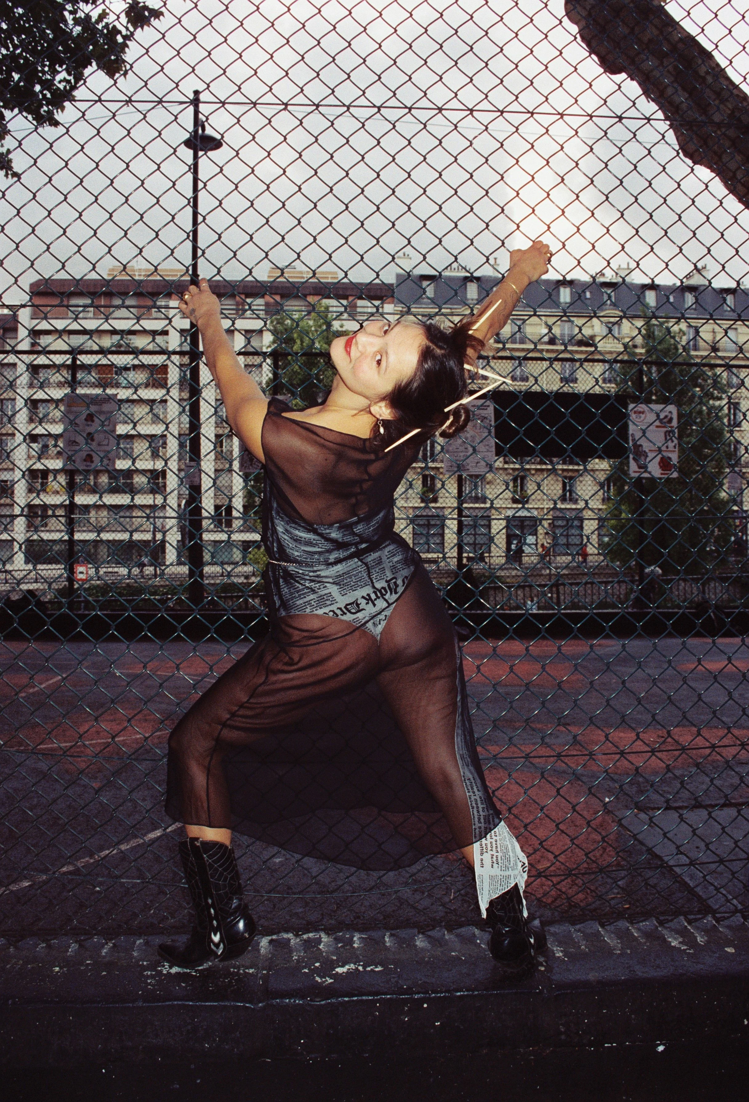 A woman in a sheer black dress with newspaper print details, black tights, and cowboy boots is standing in front of a chain-link fence, stretching her arms upward and smiling. The background shows a cityscape with buildings and a slightly overcast sky.