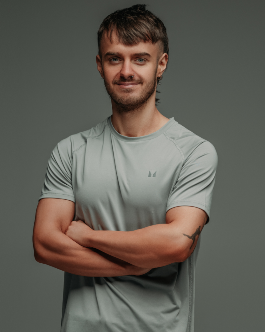 A young man with short brown hair, blue eyes, and a nose piercing, wearing a light gray athletic shirt with arms crossed, smiling slightly, with a tattoo on his left arm, in front of a gray background.