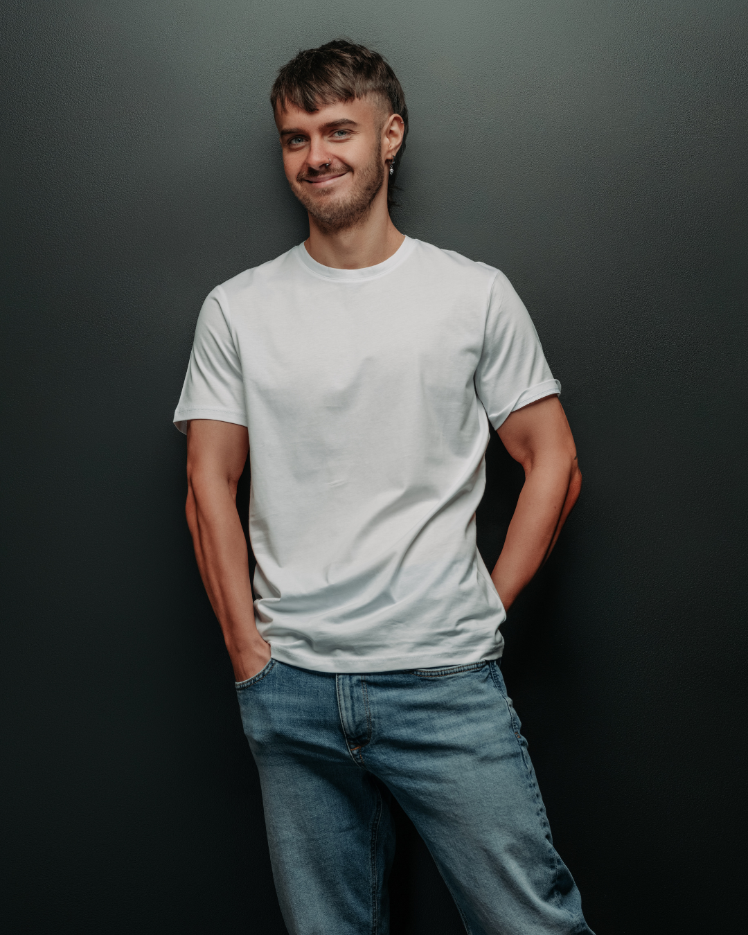 Portrait of a young man with short brown hair, wearing a white t-shirt and jeans, standing against a dark wall, smiling with hands in pockets.