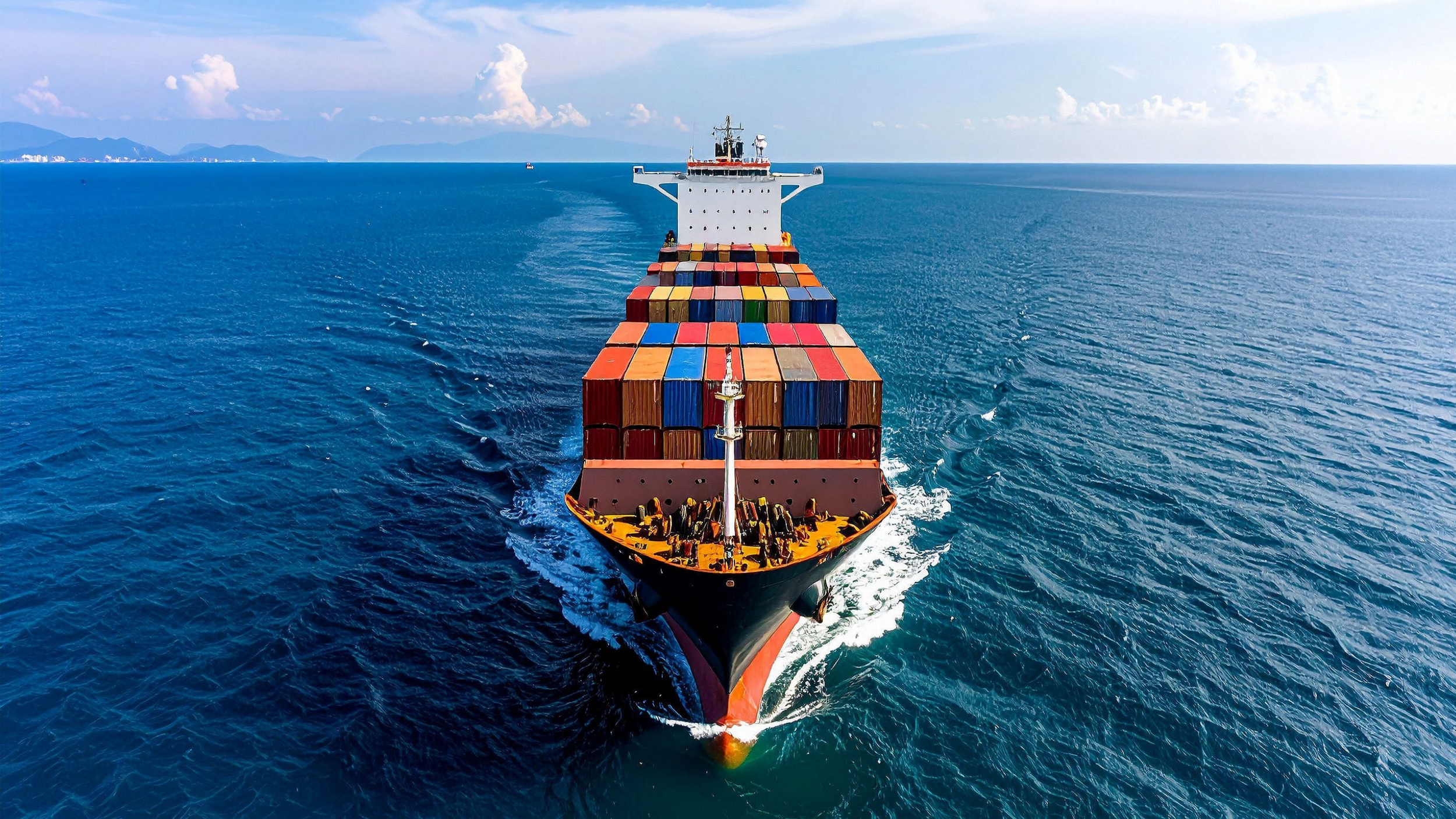 A large cargo ship carrying colorful shipping containers is sailing through the ocean with a distant coastline and mountains on the horizon.