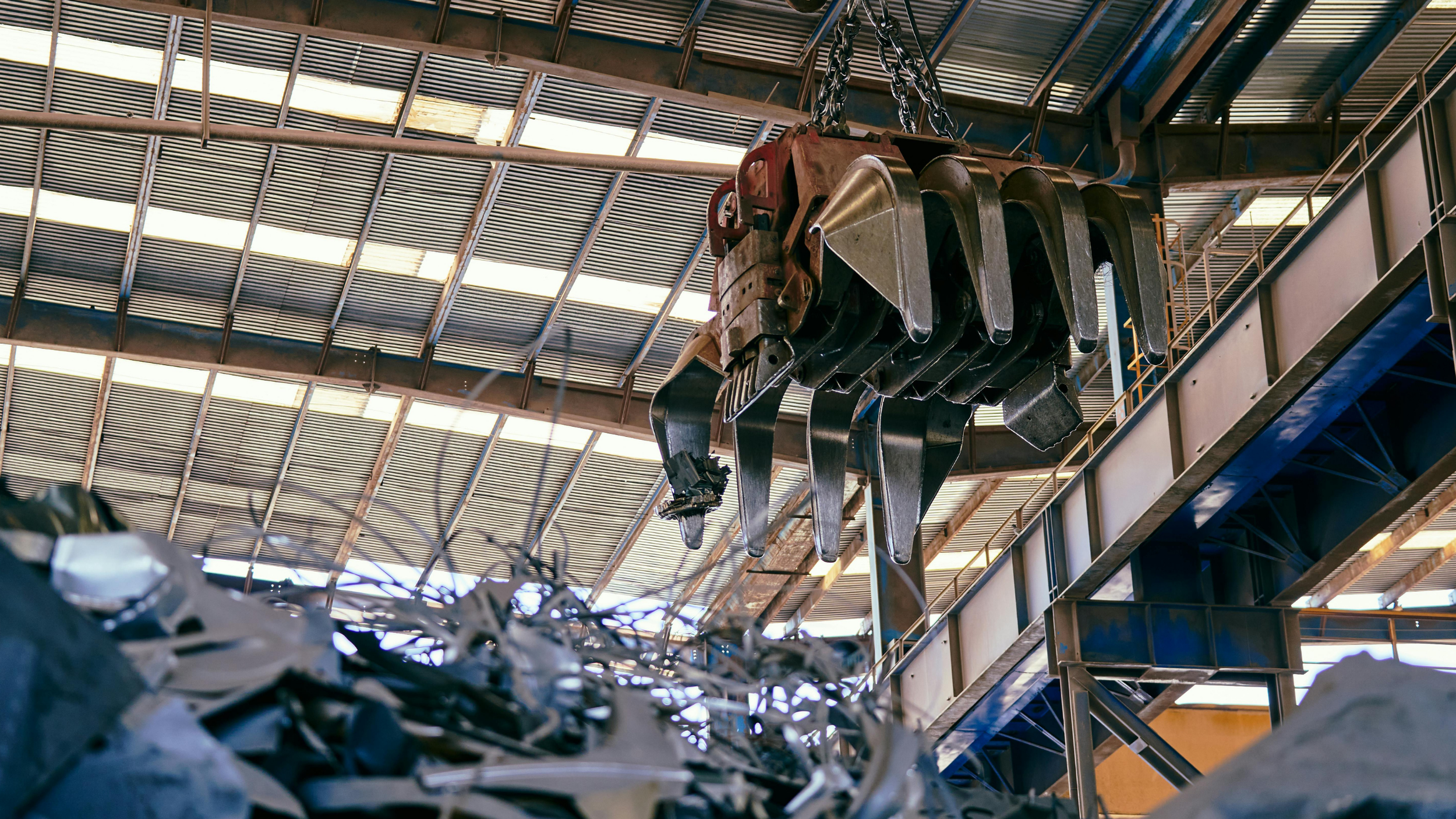 A large industrial crane with metallic claws hanging from chains inside a warehouse with metal roofing and beams.