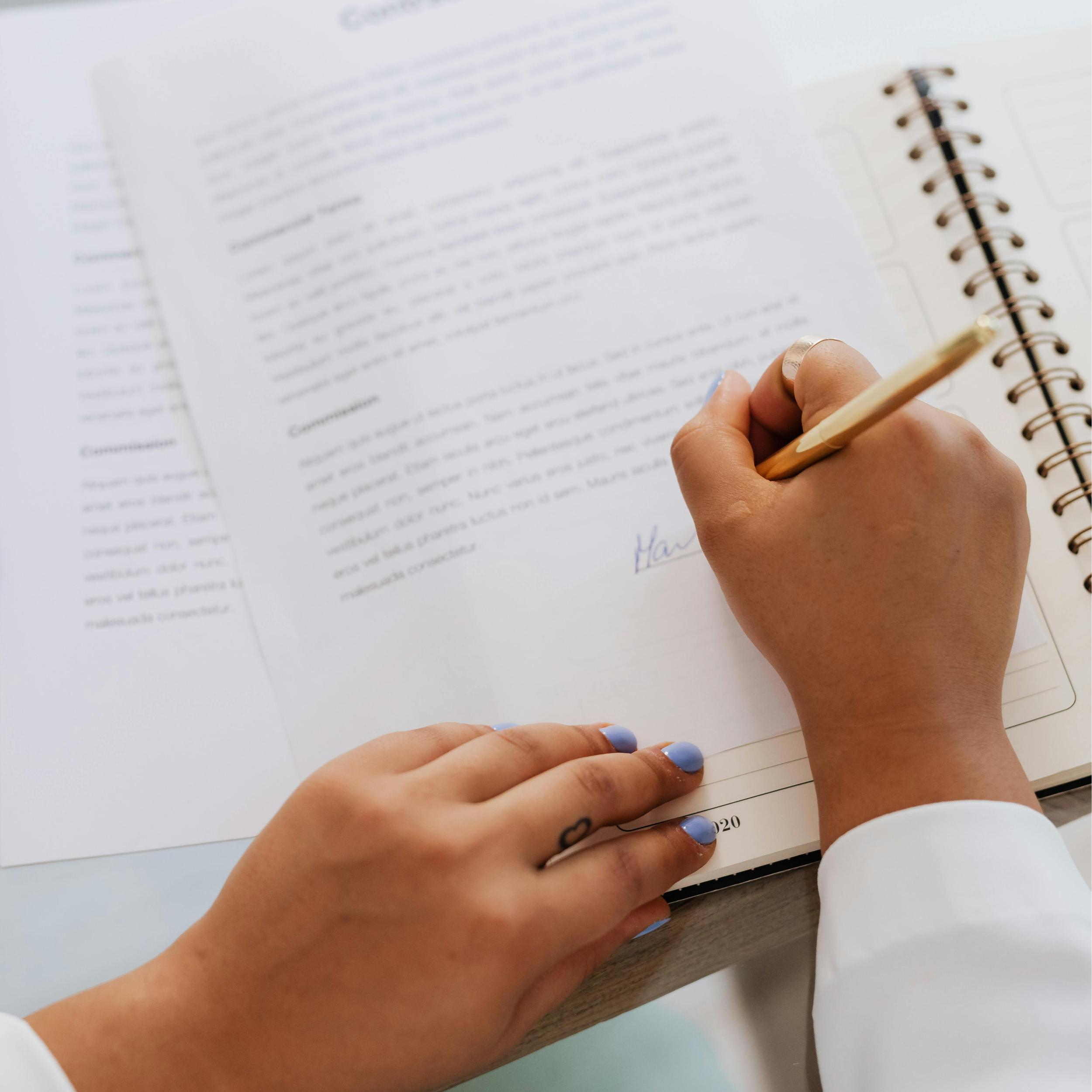 Close-up of a person with blue nail polish holding a wooden pencil, writing in a spiral notebook with white pages and printed text, on a white table.