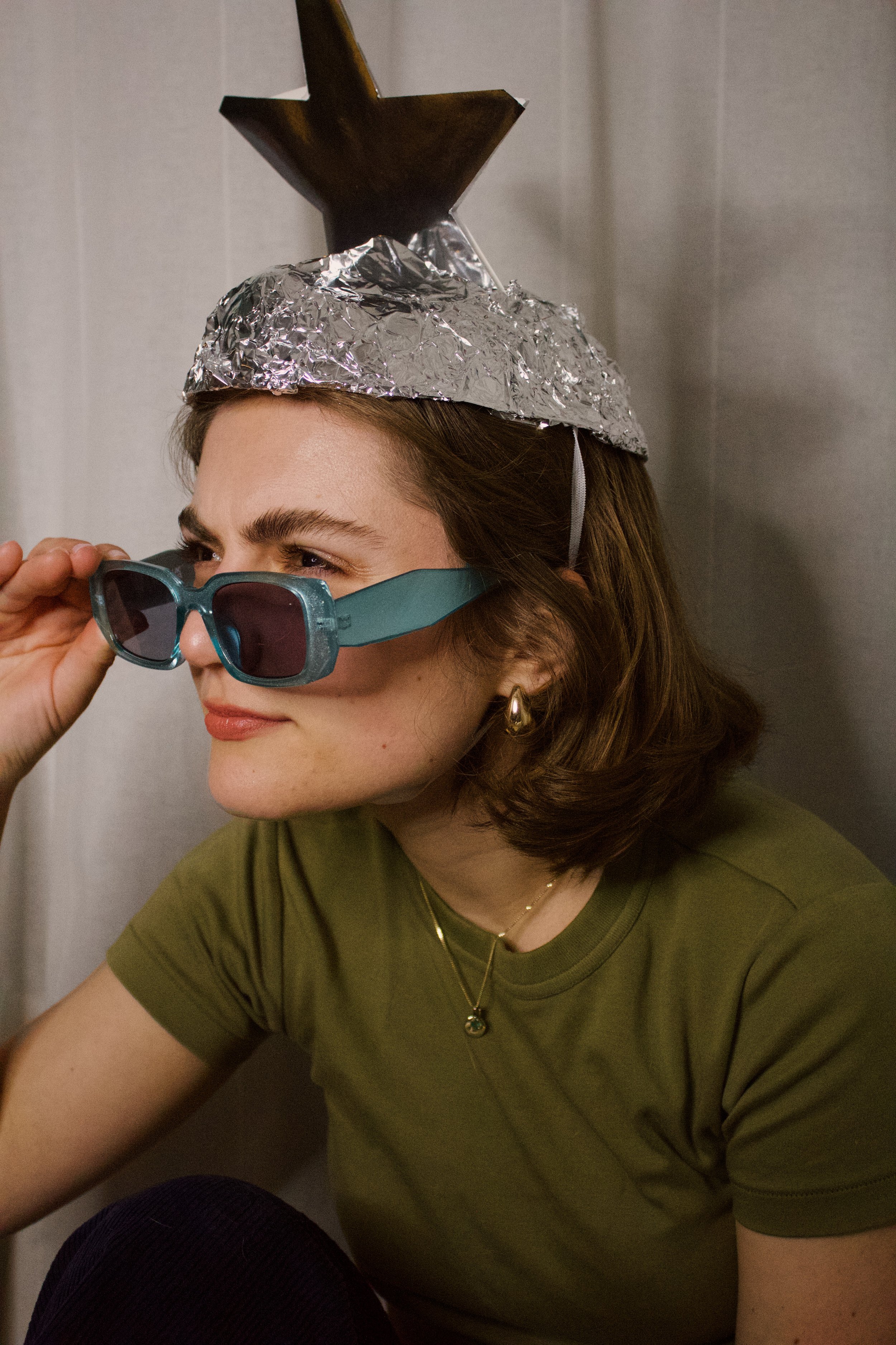 A woman with brown hair, wearing teal sunglasses, gold earrings, a green shirt, and a gold necklace, is sitting and adjusting her sunglasses. She has a foil party hat with a gold star on top on her head.