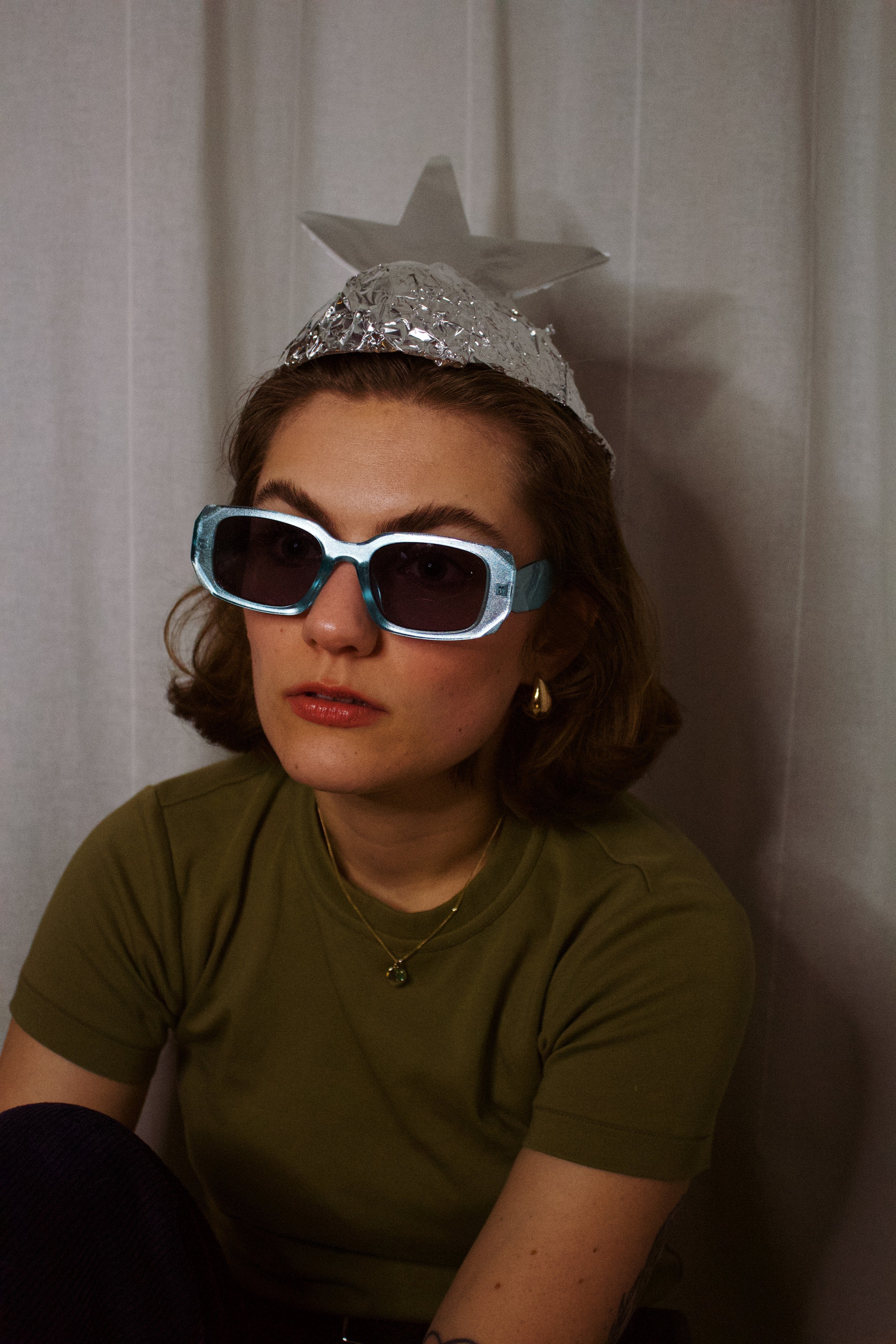 A woman wearing large sunglasses, a metallic star-shaped headband, and a green shirt, sitting against a plain wall.